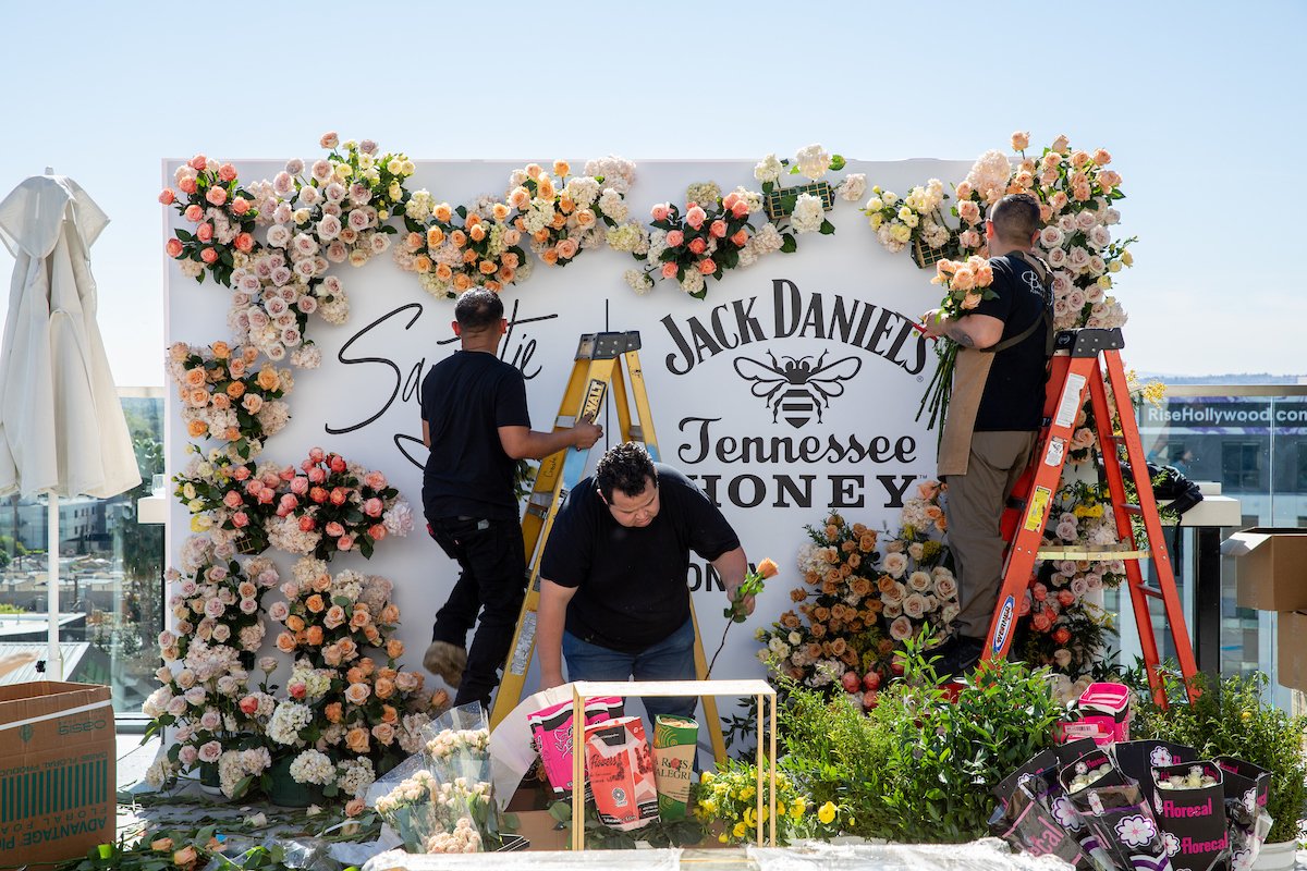 People decorating a floral wall backdrop with roses and other flowers for an event, with a Jack Daniel's and Tennessee Honey logo.