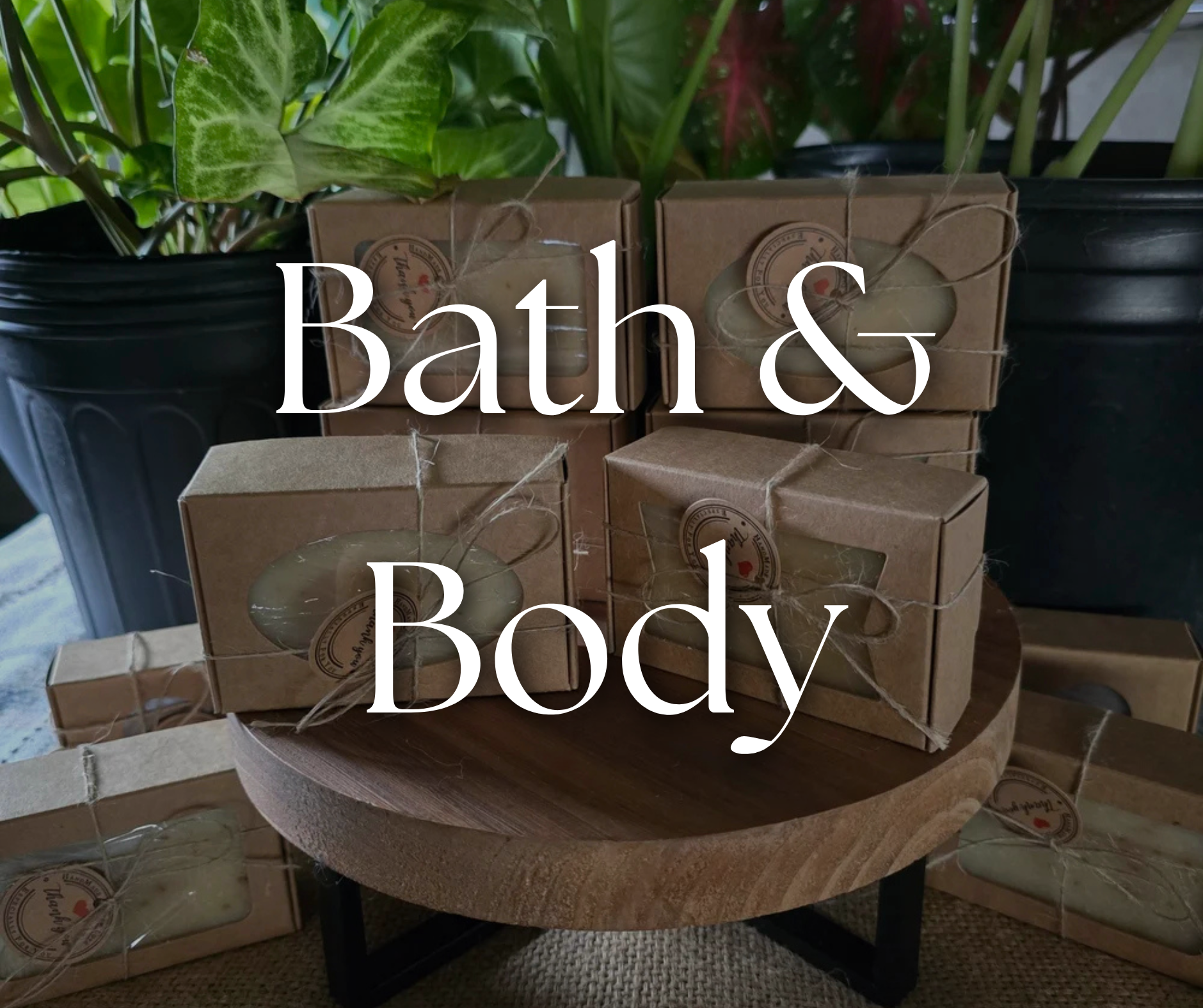 Various bath and body products in brown packaging with clear windows, displayed on a small wooden table, with potted plants in the background.
