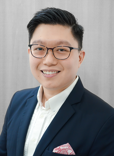 A young man dressed in a dark suit jacket and white dress shirt, smiling, with glasses, against a plain gray background.