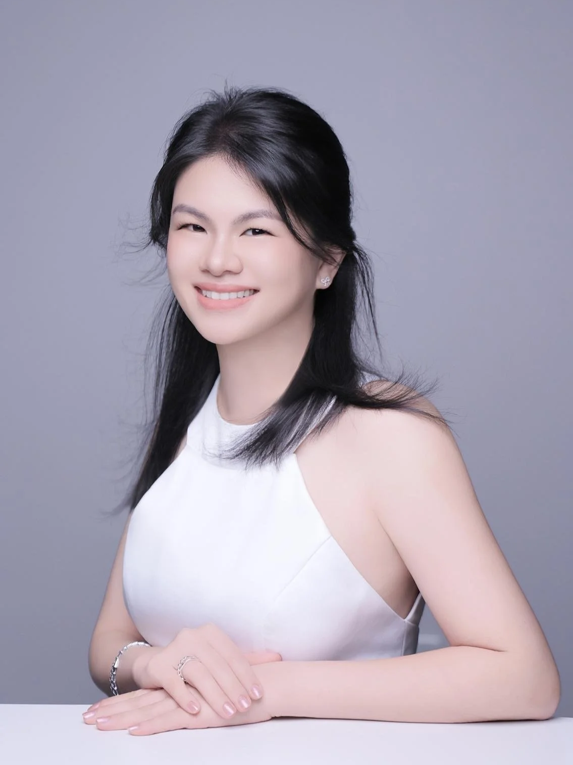 A woman with long black hair, glasses, and earrings smiling, wearing a dark floral dress against a plain background.