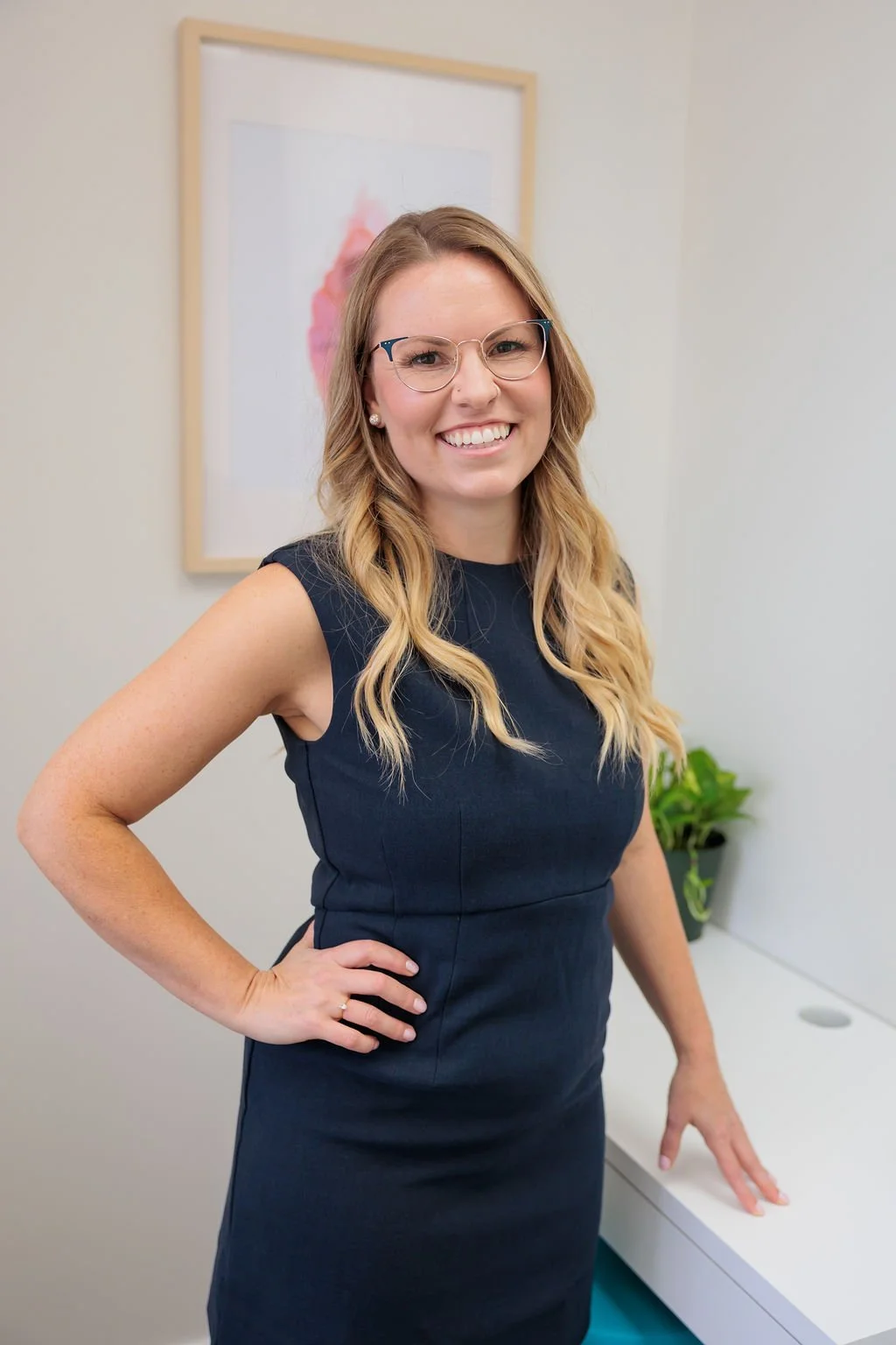 A woman with glasses and long wavy hair smiling, dressed in a navy sleeveless dress, standing in an office space with a framed picture on the wall and a green plant on a white table.