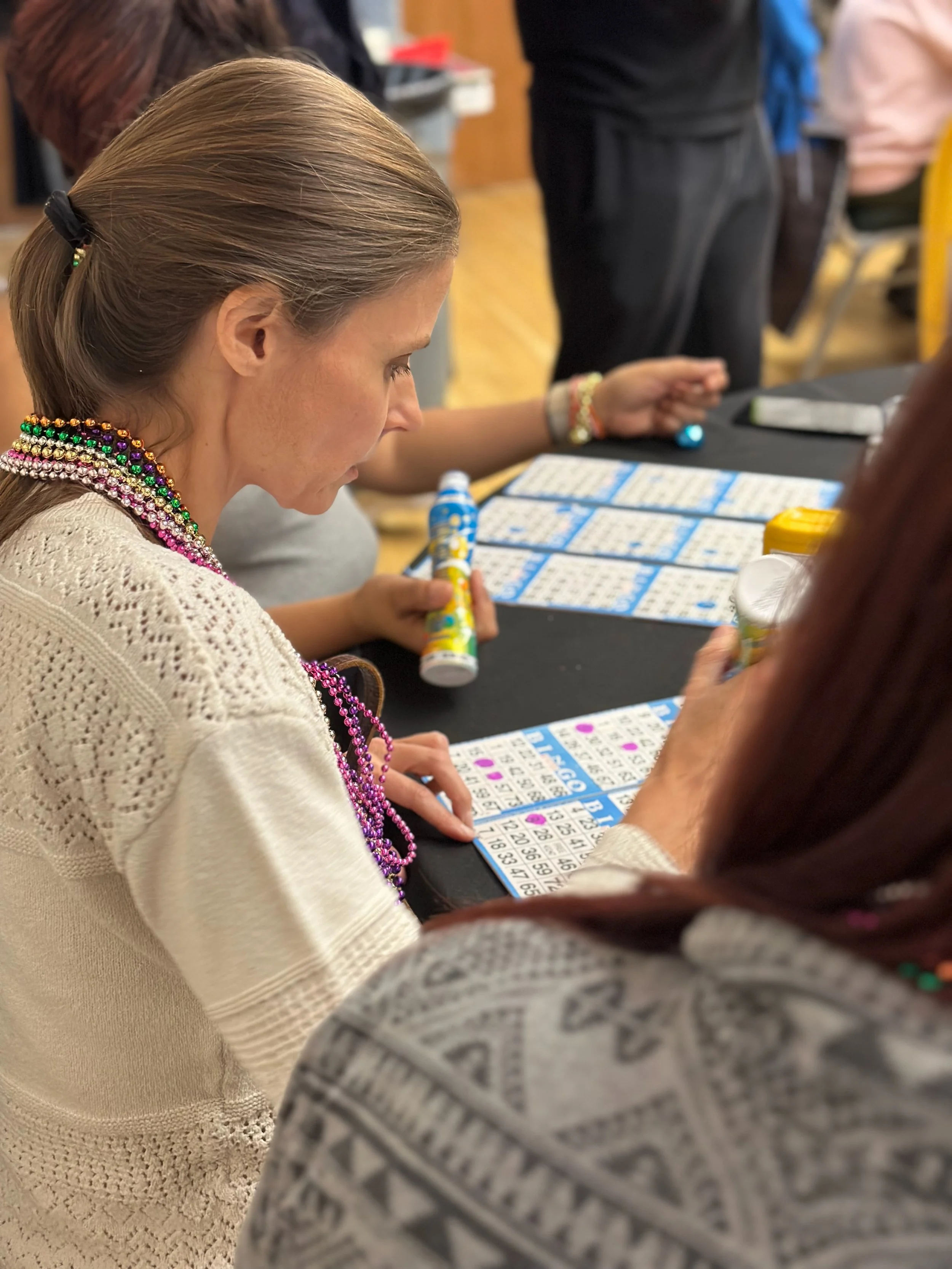 Participant wearing colorful bead necklaces concentrates on a bingo card while holding a bingo dauber at a community event table.