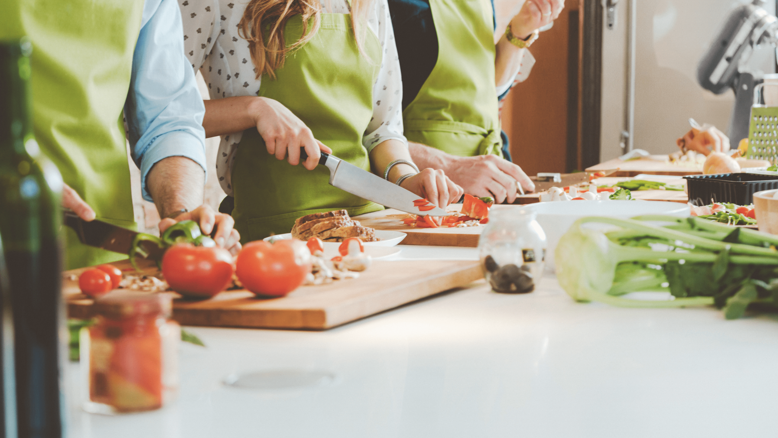 Close-up of several people wearing green aprons preparing food, slicing tomatoes, peppers, and bread on wooden cutting boards in a shared kitchen space.