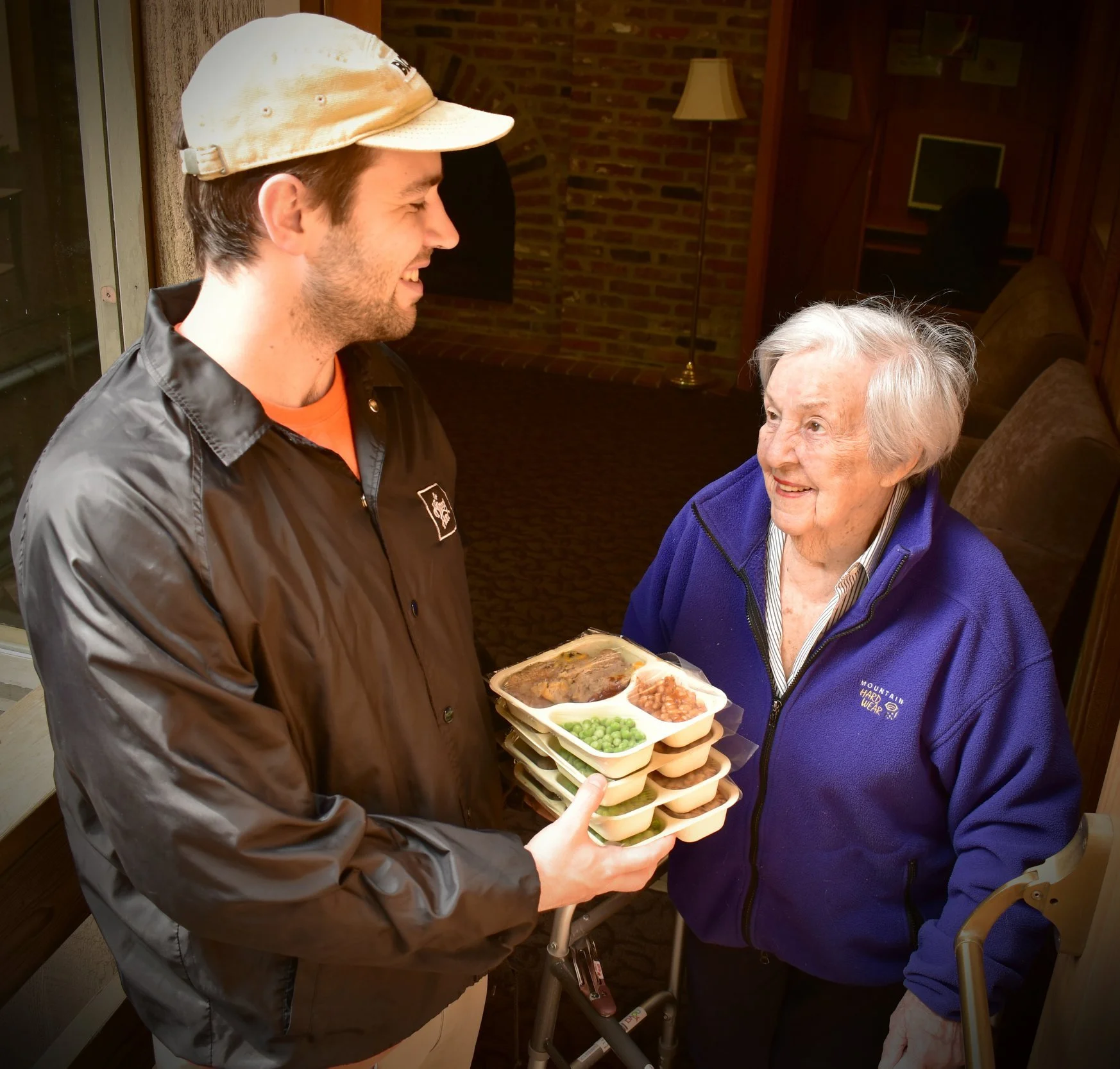 A volunteer hands stacked meal containers to a smiling elderly woman during a home meal delivery.