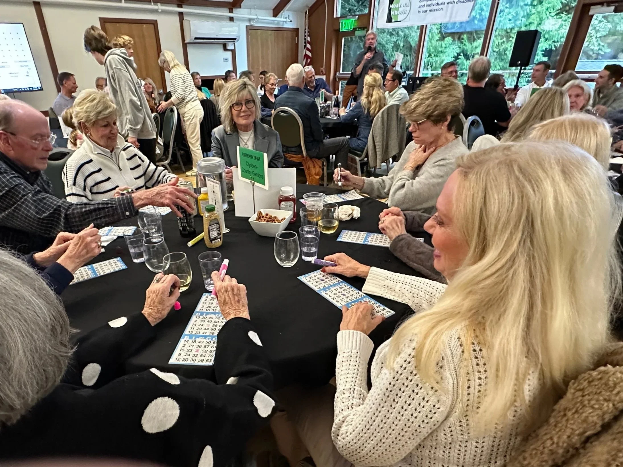 Attendees gathered around a table playing bingo at a Meals on Wheels fundraiser, holding bingo cards and markers.
