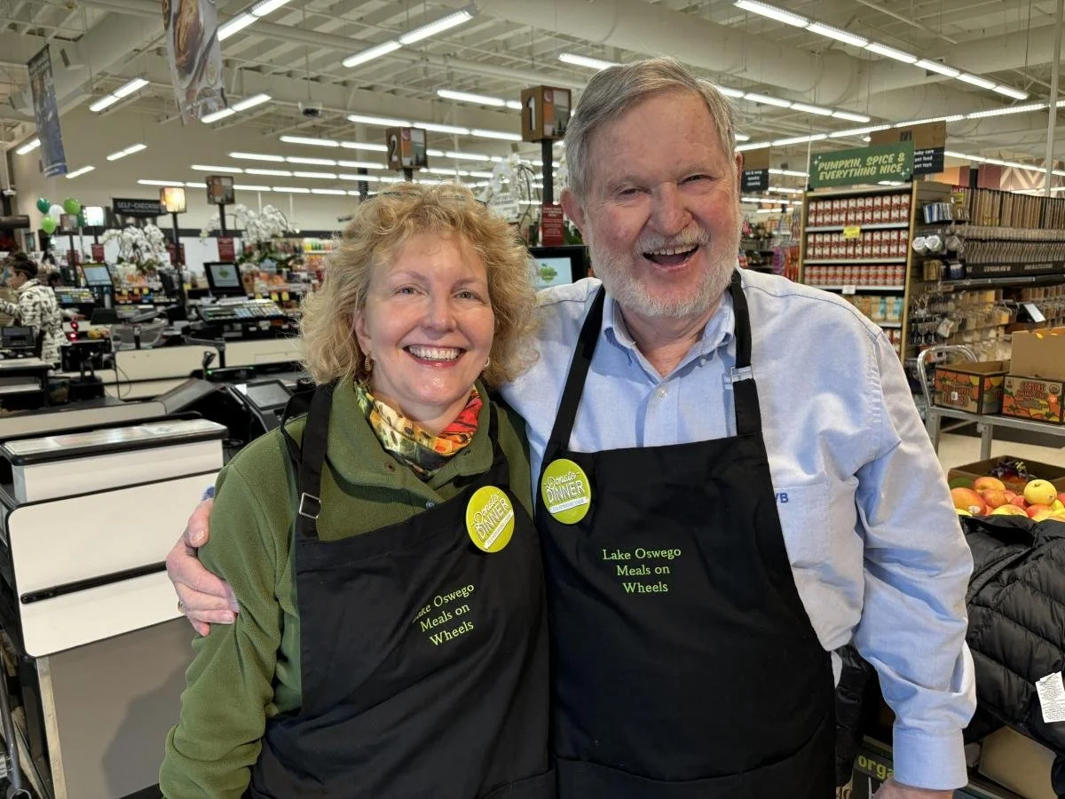 Two smiling volunteers wearing black Lake Oswego Meals on Wheels aprons stand together inside a grocery store during a community fundraising event.