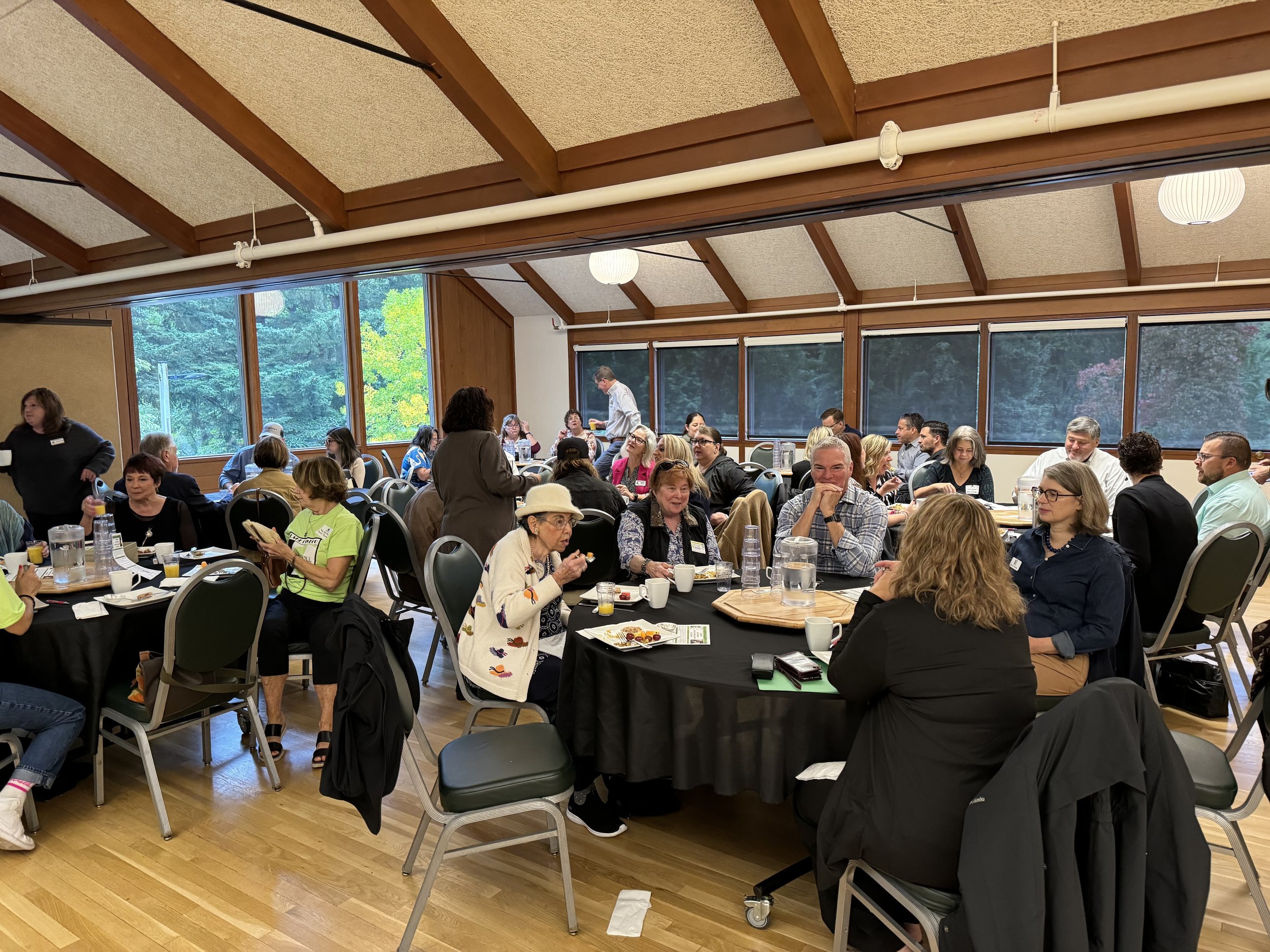 Community members seated at round tables during a Meals on Wheels event, listening to a speaker in a bright room with large windows.