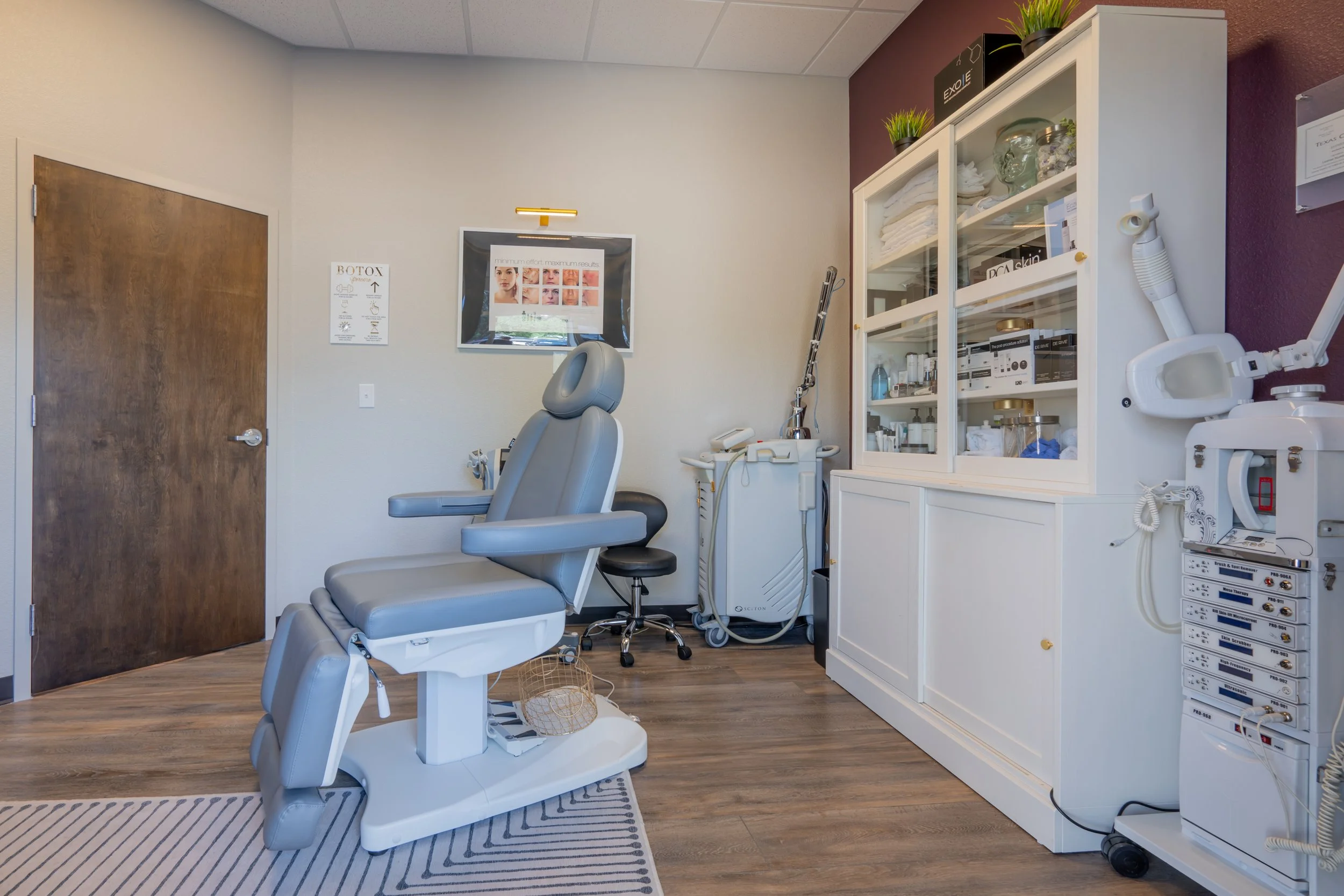A medical treatment room with a reclining chair, medical equipment, and a white cabinet with supplies, decorated with green plants on top.