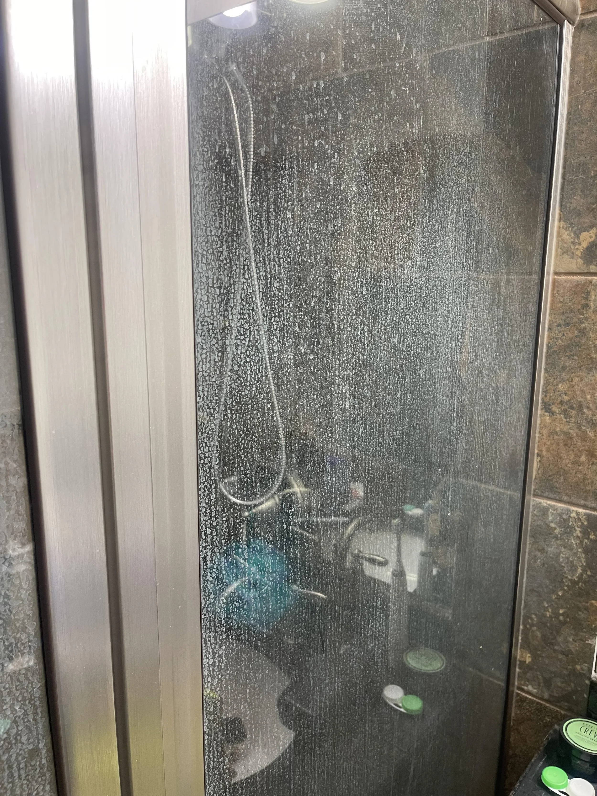 Shower with a glass door showing signs of mineral buildup, with a handheld showerhead.