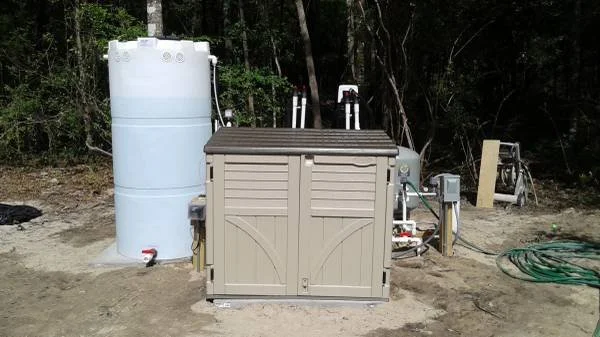 An outdoor reverse osmosis water treatment system to remove rotten egg smell, hardness, stains, bacteria and discoloration with a pressure tank, and plumbing components installed on a concrete pad against a beige wall.