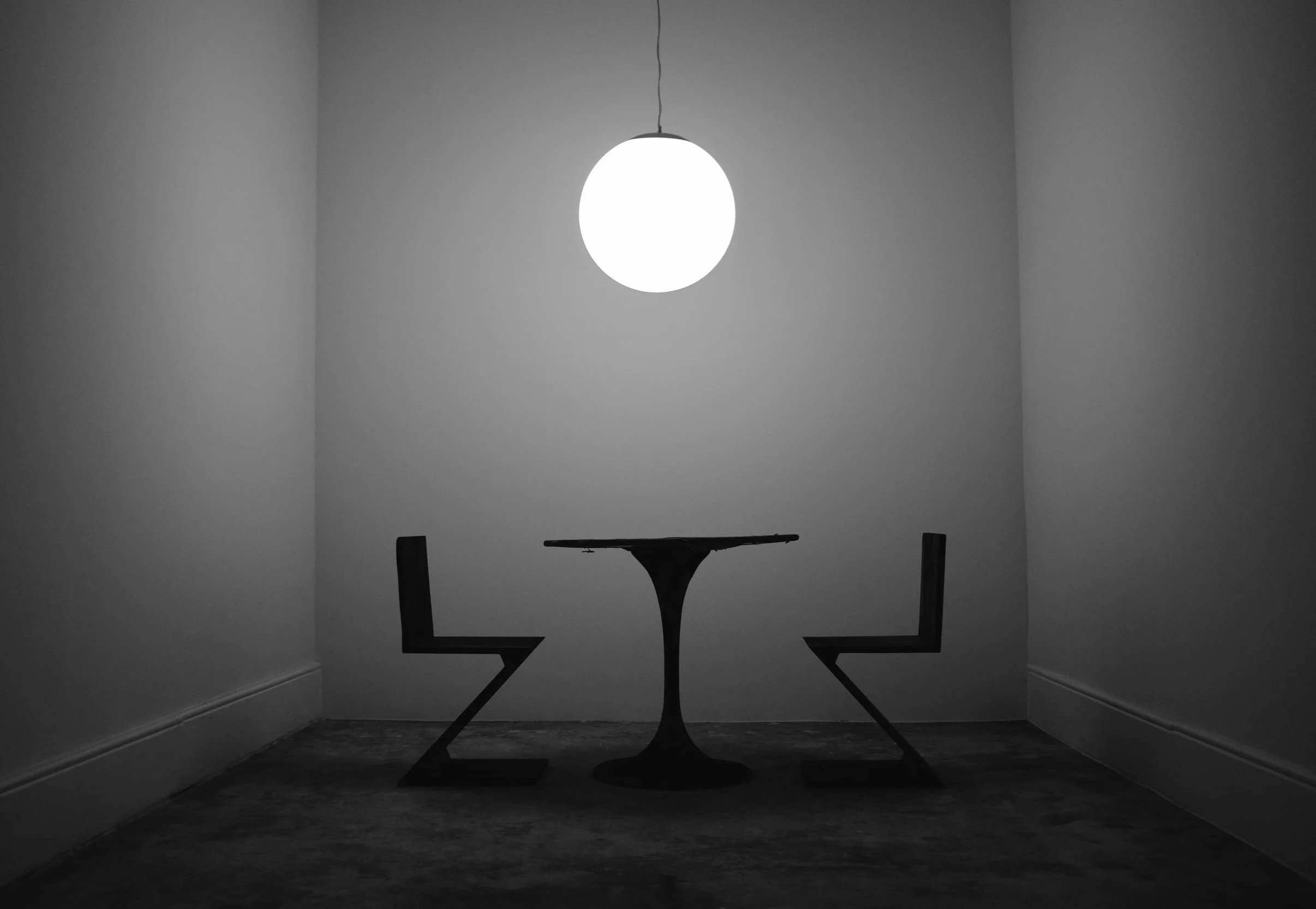 A black-and-white photo of a small table with two chairs in a corner, illuminated by a single round ceiling light at Studio Rationale.