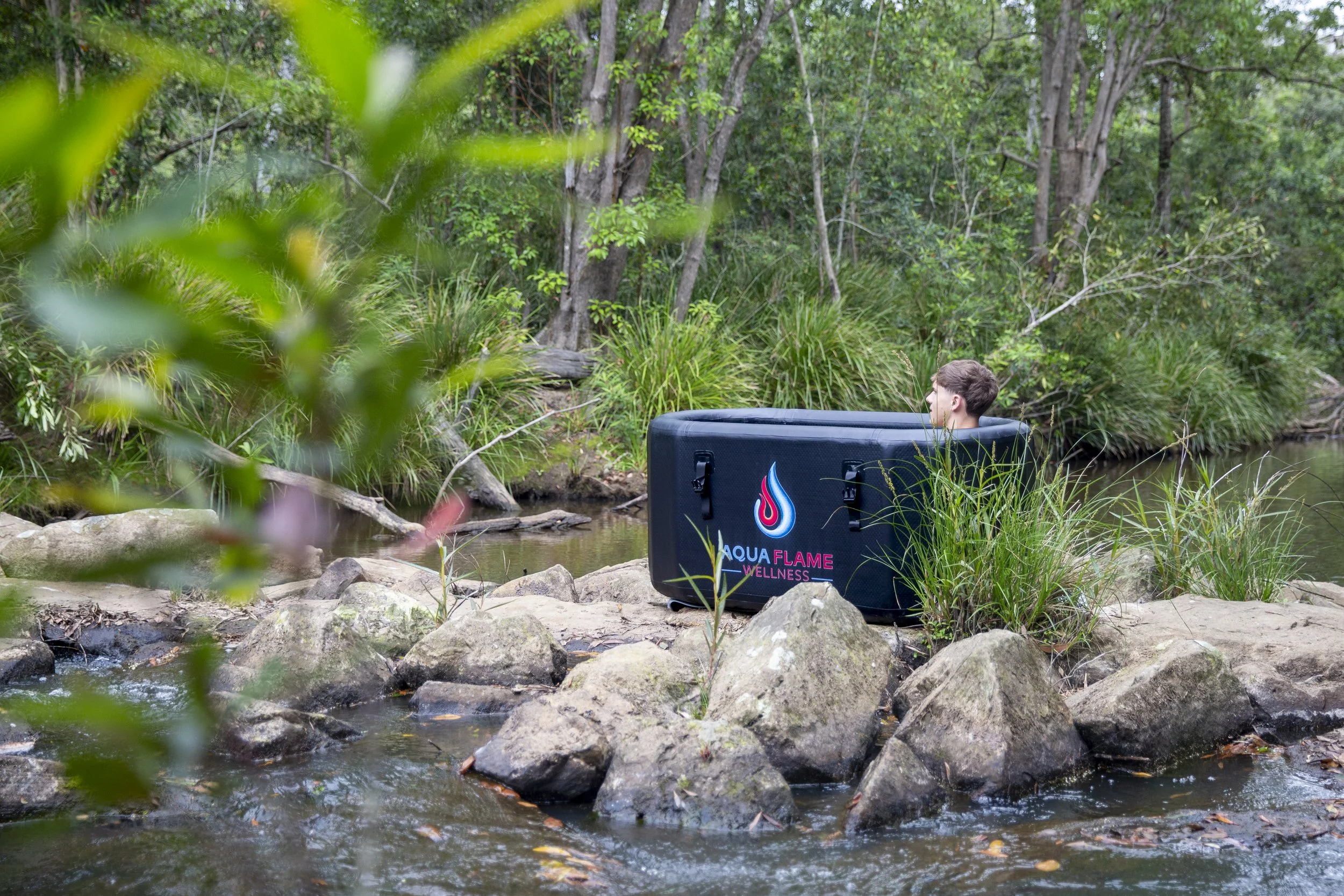 A person sitting on rocks by a creek in a forest, next to a black wellness pod with the logo and words 'Aqua Flame Wellness' on it.