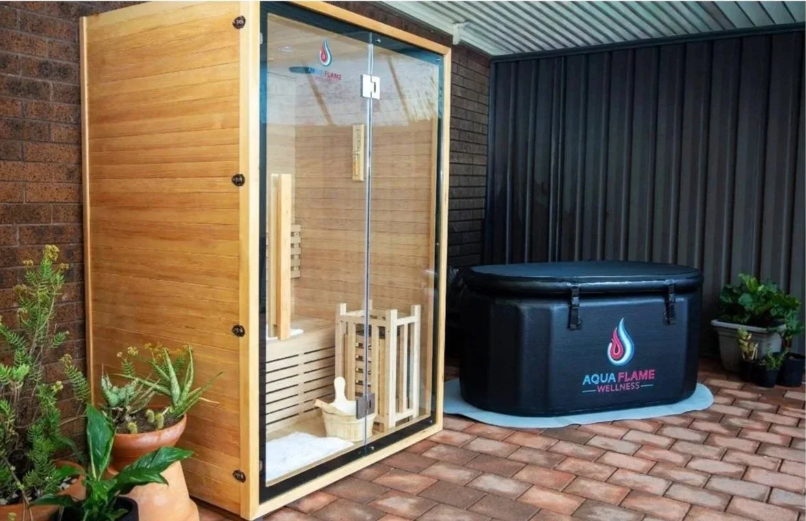 An outdoor wellness setup with a wooden sauna and a black hot tub labeled 'Aqua Flame Wellness' on a brick patio, with potted plants nearby.