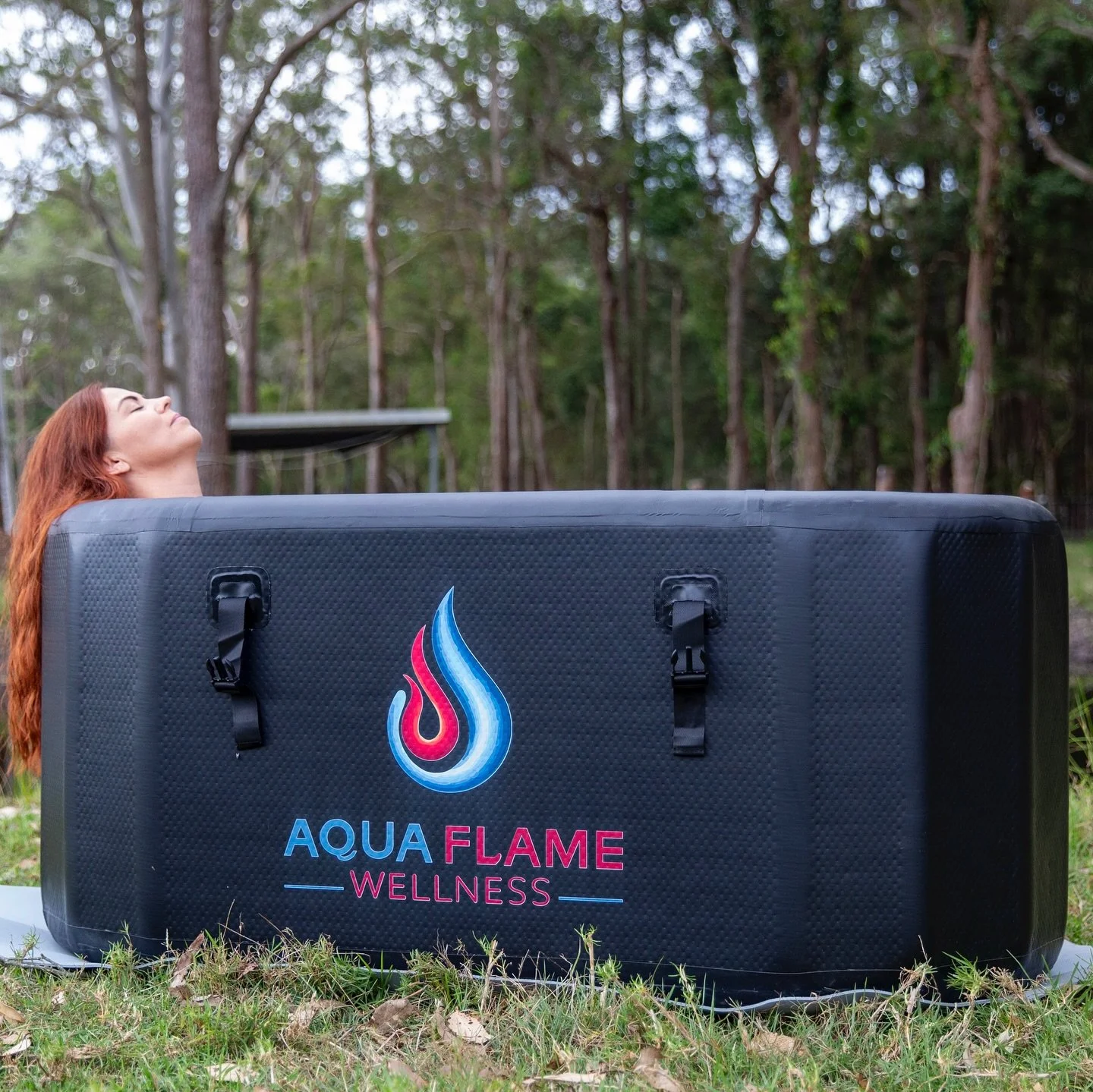 A woman relaxing with her eyes closed behind a large black wellness pod with a colorful logo and the words 'Aqua Flame Wellness' on it, outdoors in a wooded area.