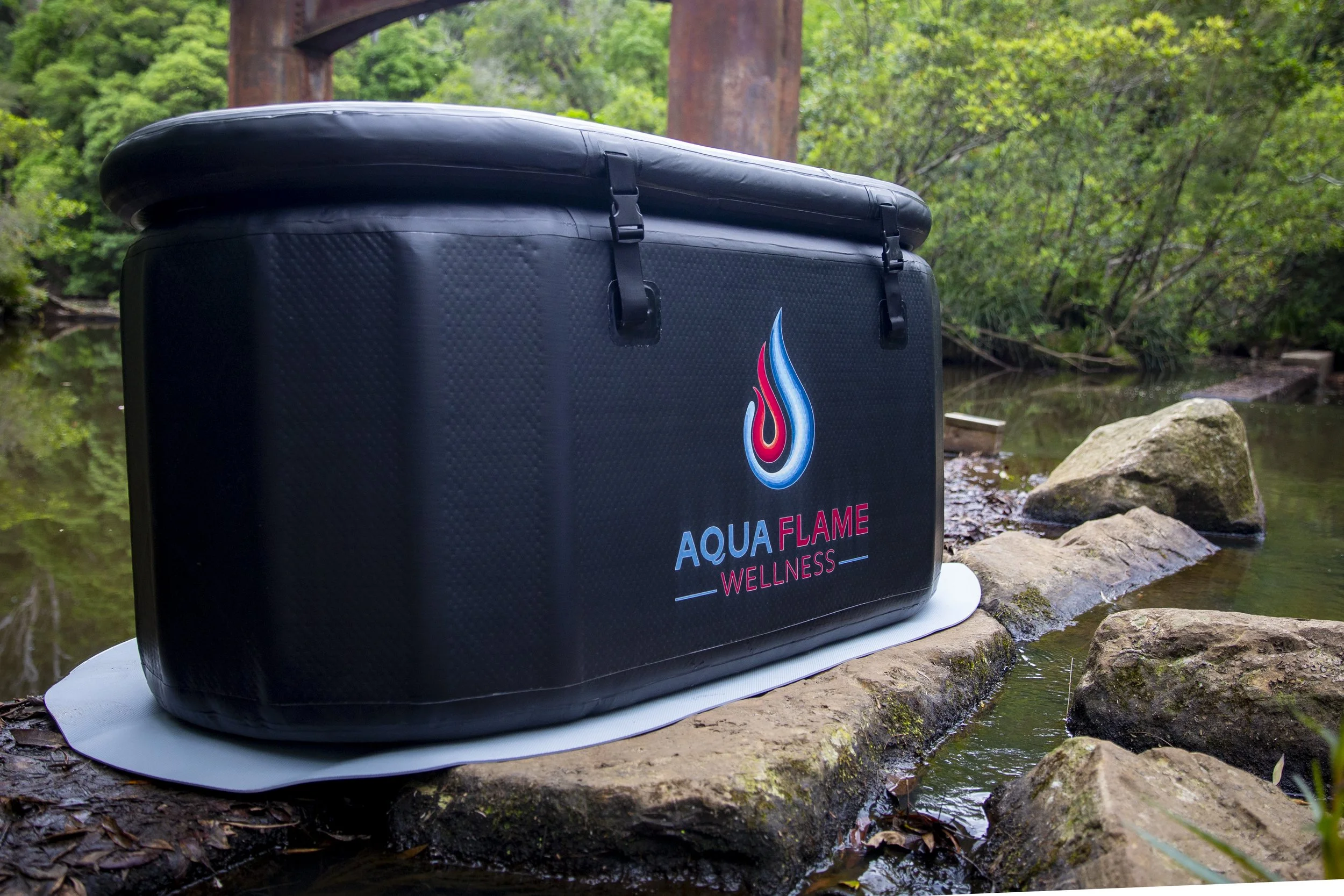 Black waterproof wellness box with 'Aqua Flame Wellness' logo placed on a rock near a small stream with rocks and green trees in background.