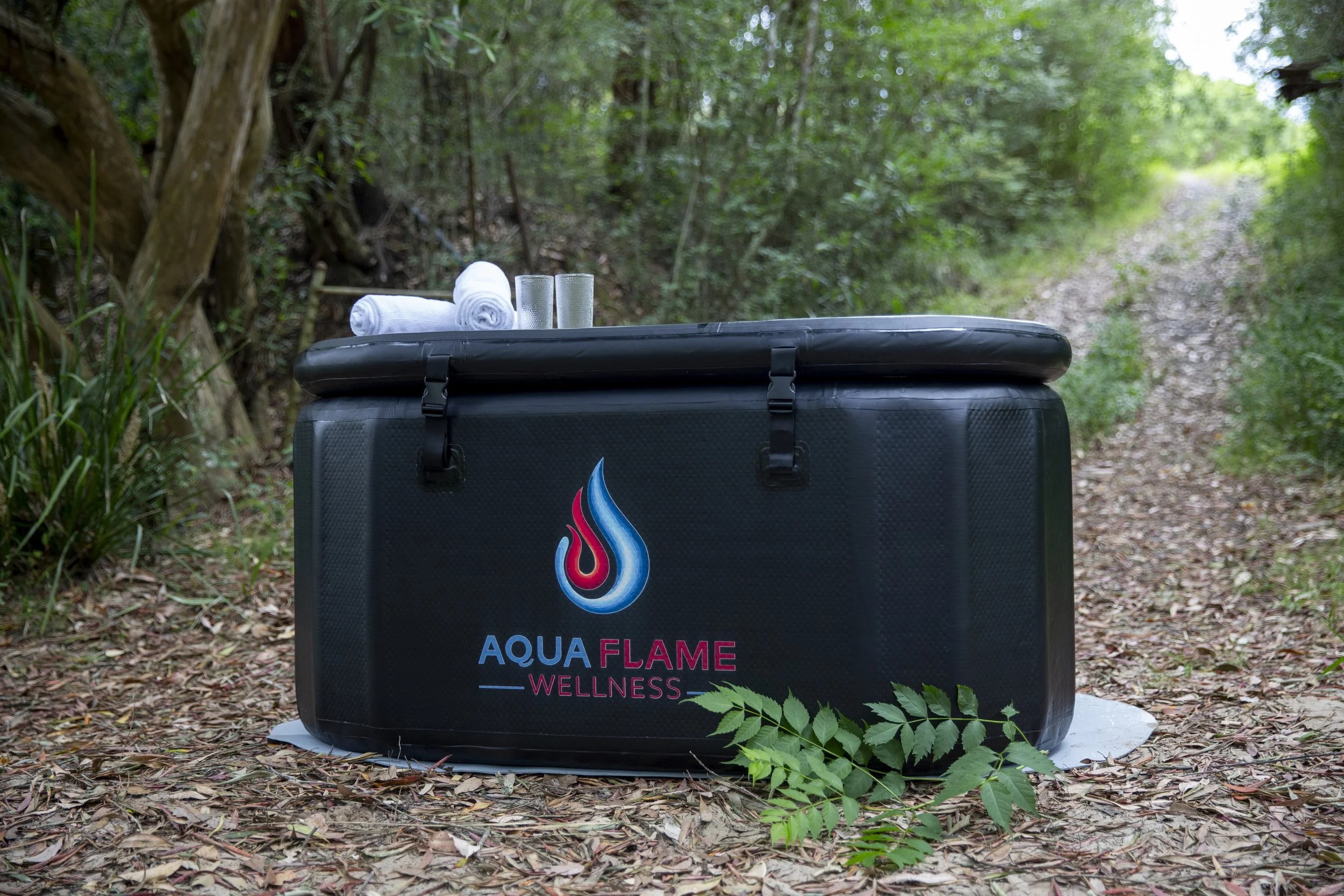 A portable wellness spa ice bath with the logo 'Aqua Flame Wellness' on it, placed outdoors on a forest trail. On top of the spa, there are two rolled-up white towels and two glasses, with lush green foliage in the background. portable ice plunge tub