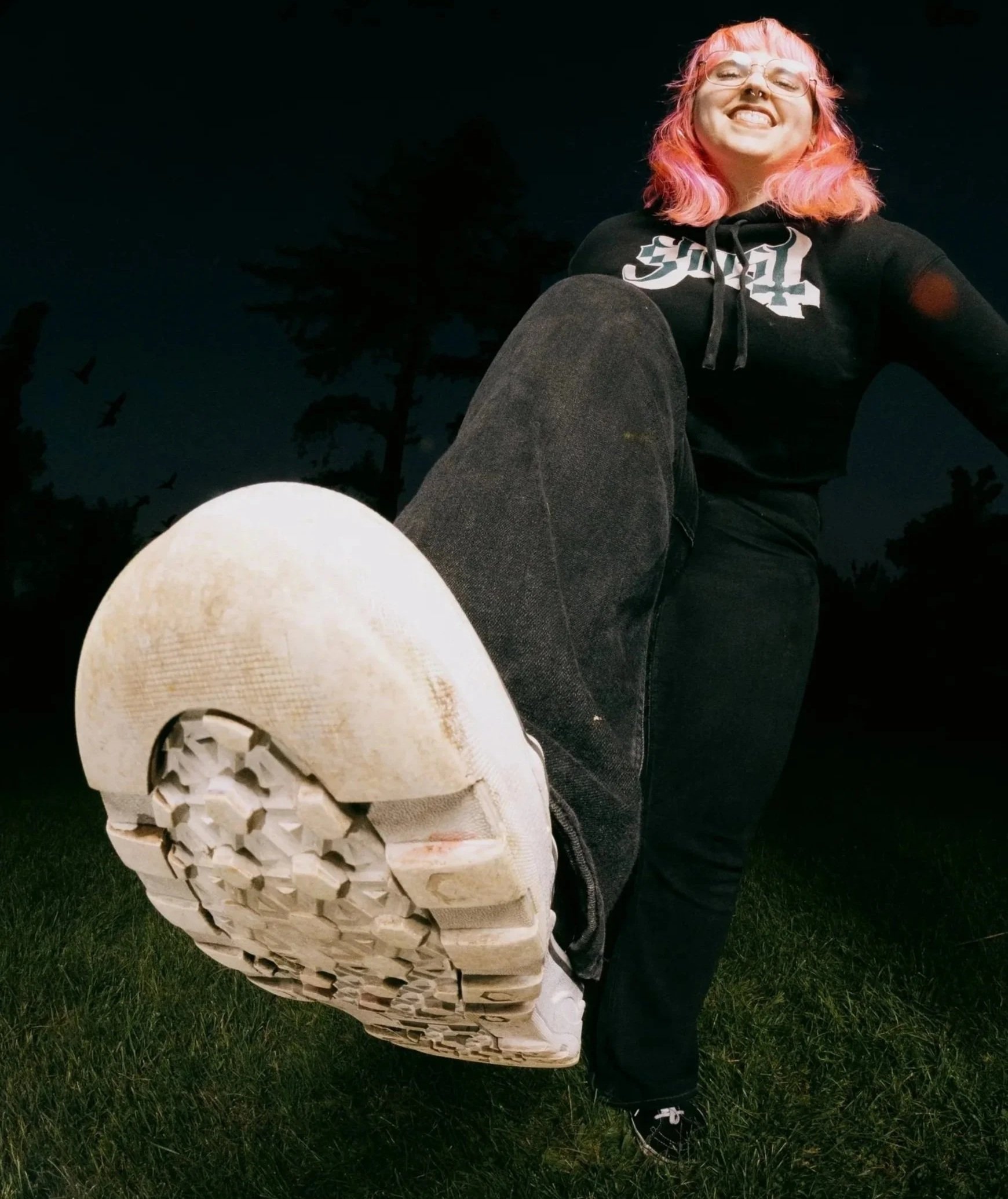 A picture of Cierra, a white nonbinary perosn with pink hair, wearing black clothes, kicking at the camera