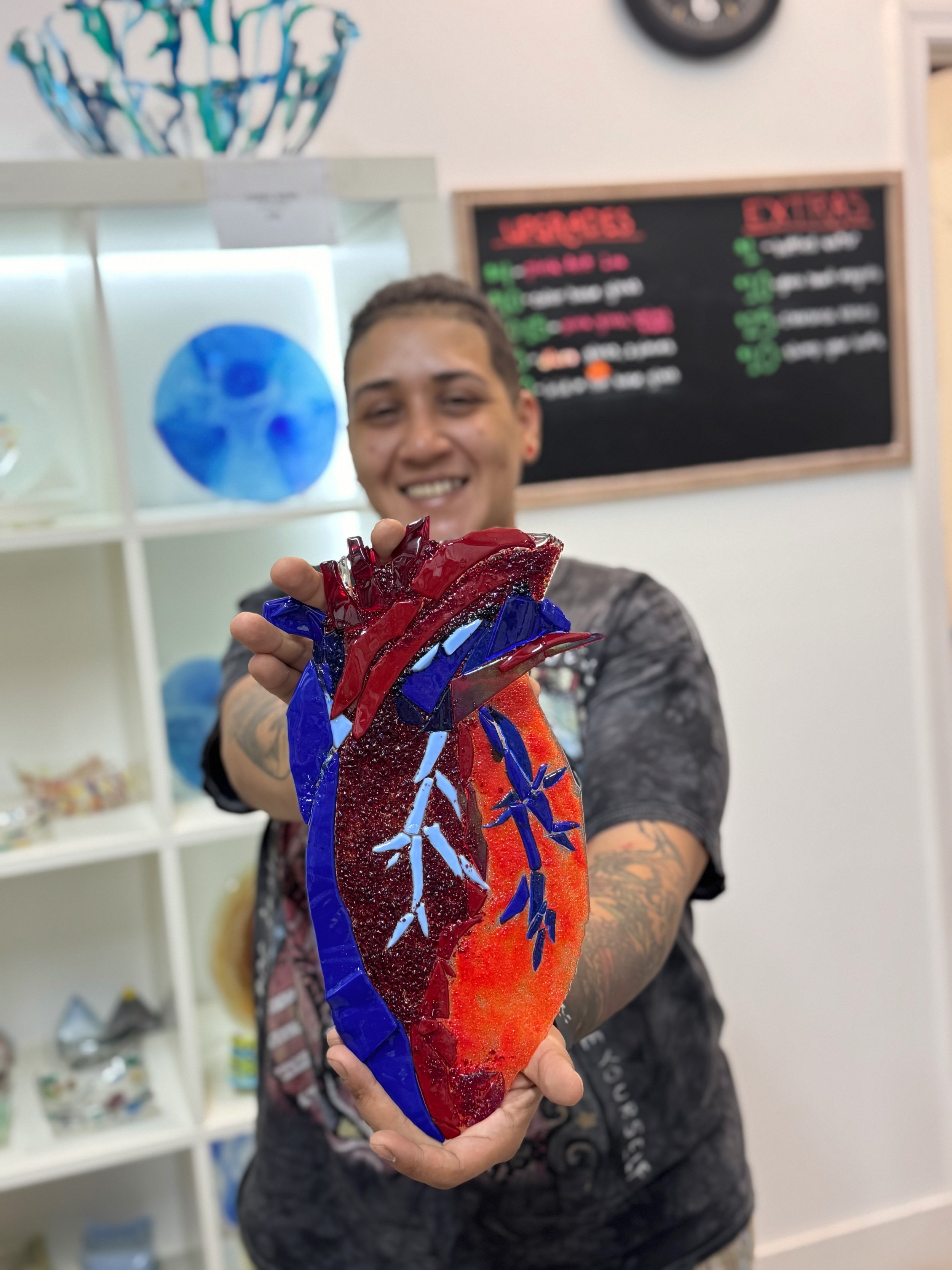 Ishiamary RC holding up a colorful glass art piece shaped like a human heart, with blue, red, and orange colors and decorative light blue branches at Sarasota School of Glass.