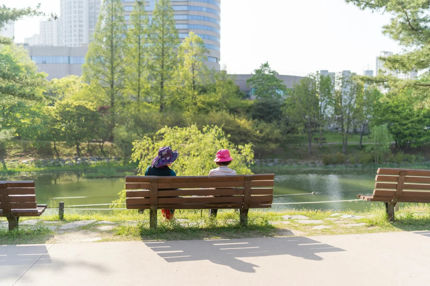 Two children sitting on a park bench near a pond, surrounded by green trees and high-rise buildings in the background.