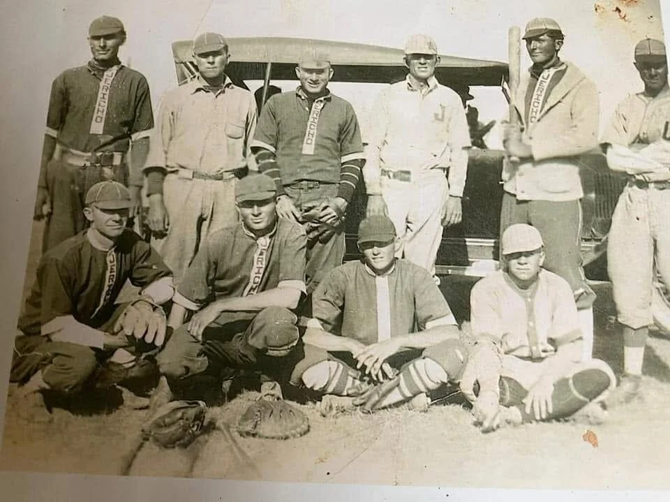 Black and white photo of a group of young boys in baseball uniforms posing outdoors in front of a truck, some sitting and some standing, with baseball gloves and equipment on the ground.