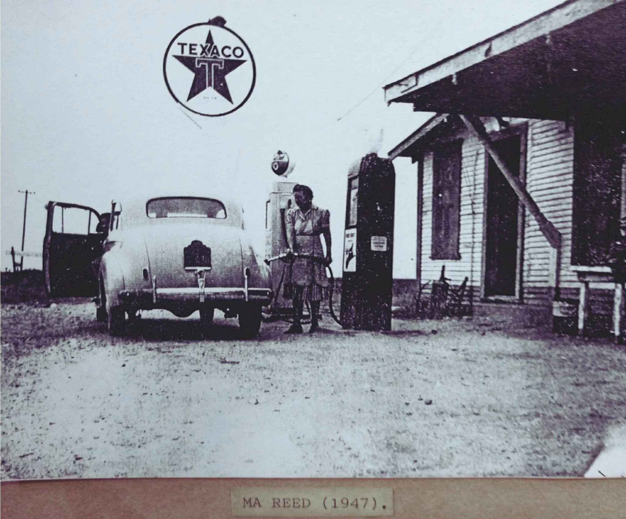 Black and white photo from 1947 showing a woman refueling a vintage car at a Texaco gas station, with a sign overhead displaying the Texaco logo.