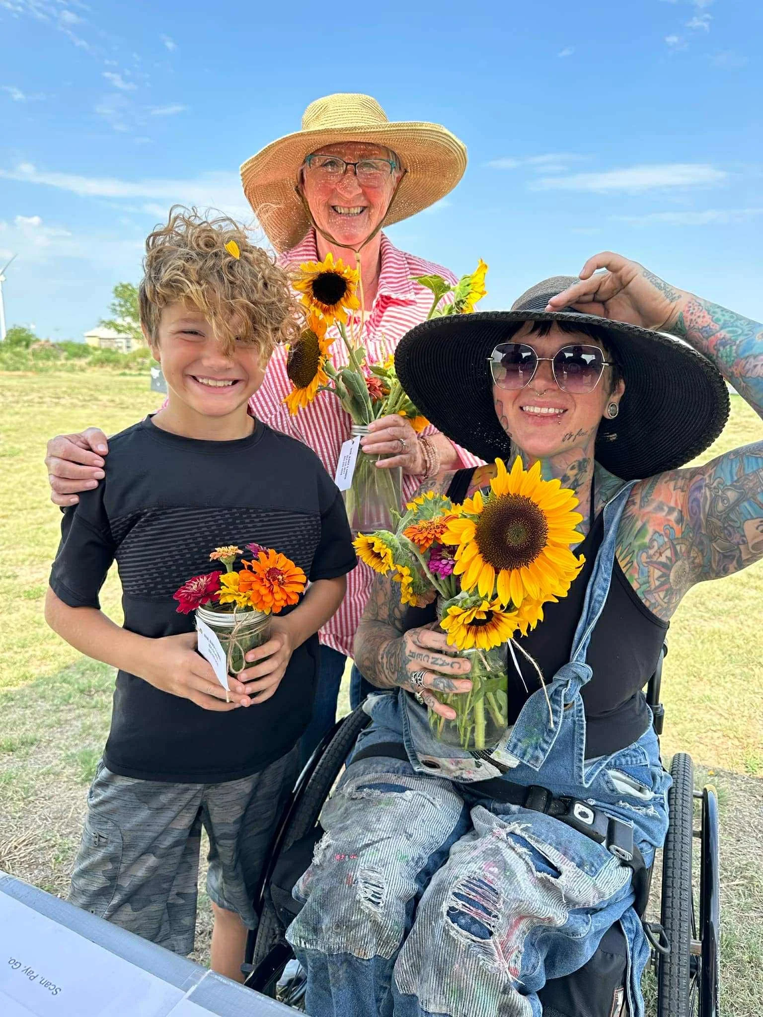 Three happy people, a young boy, a woman with tattoos wearing sunglasses and a large sun hat, and an elderly woman with a wide brim hat, holding sunflowers and smaller flowers outdoors on a sunny day.