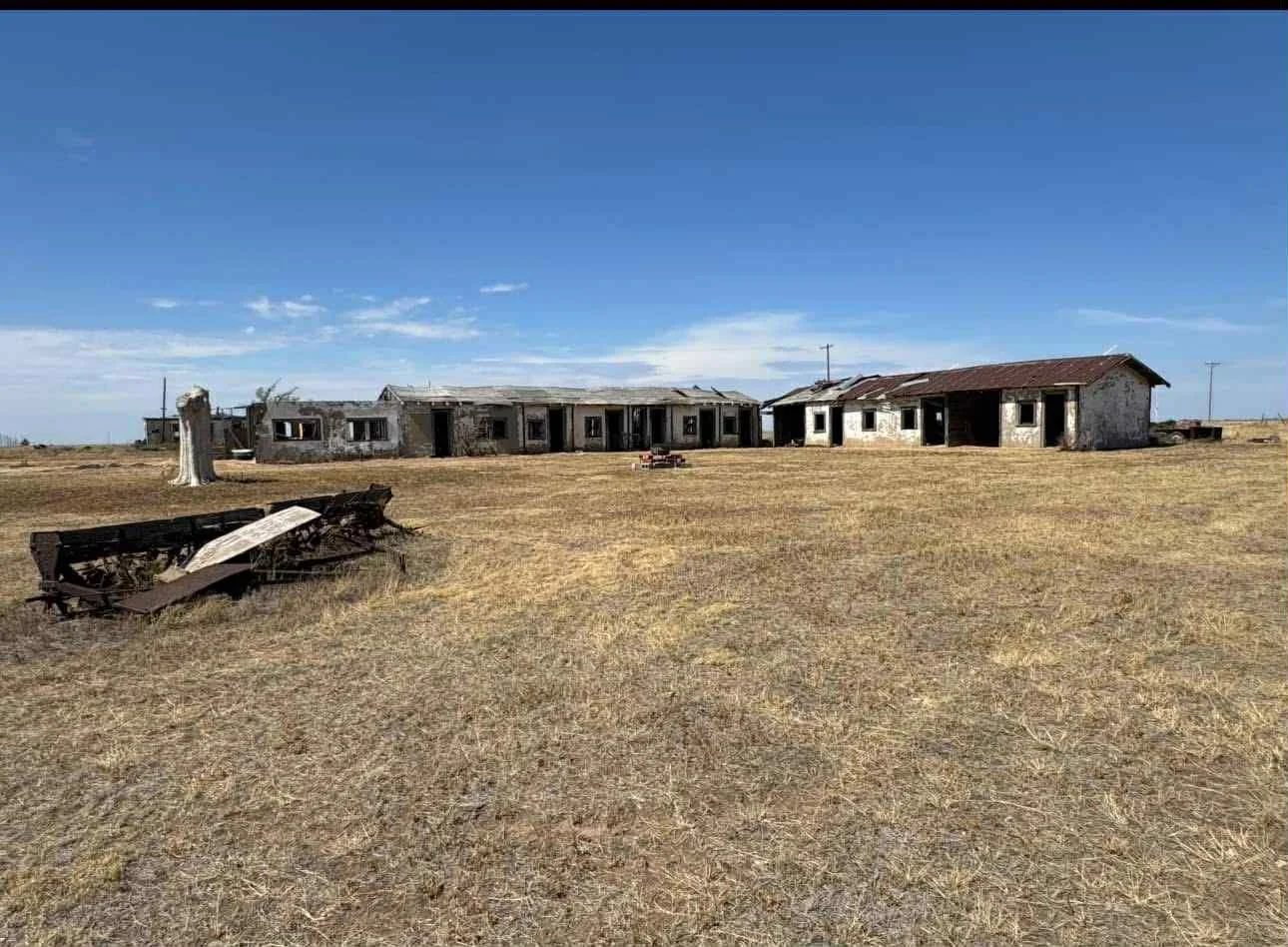 An abandoned, dilapidated farm with broken buildings and overgrown grass under a blue sky.