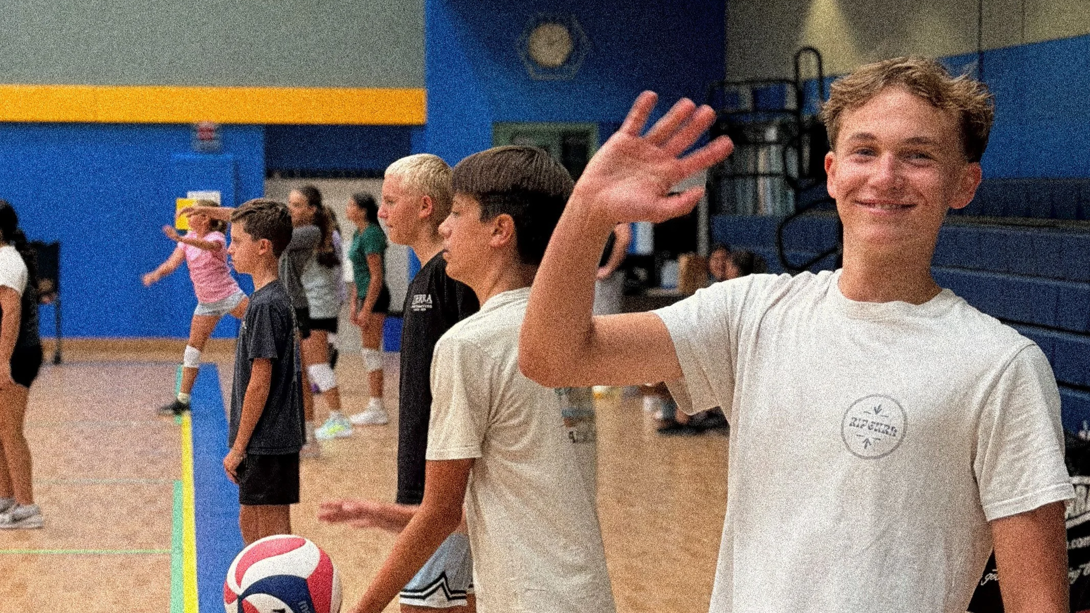 A group of children at an indoor volleyball practice, with one smiling boy in a white T-shirt waving at the camera in the foreground. Others are lined up behind him, some holding a volleyball, in a gym with blue and yellow walls.