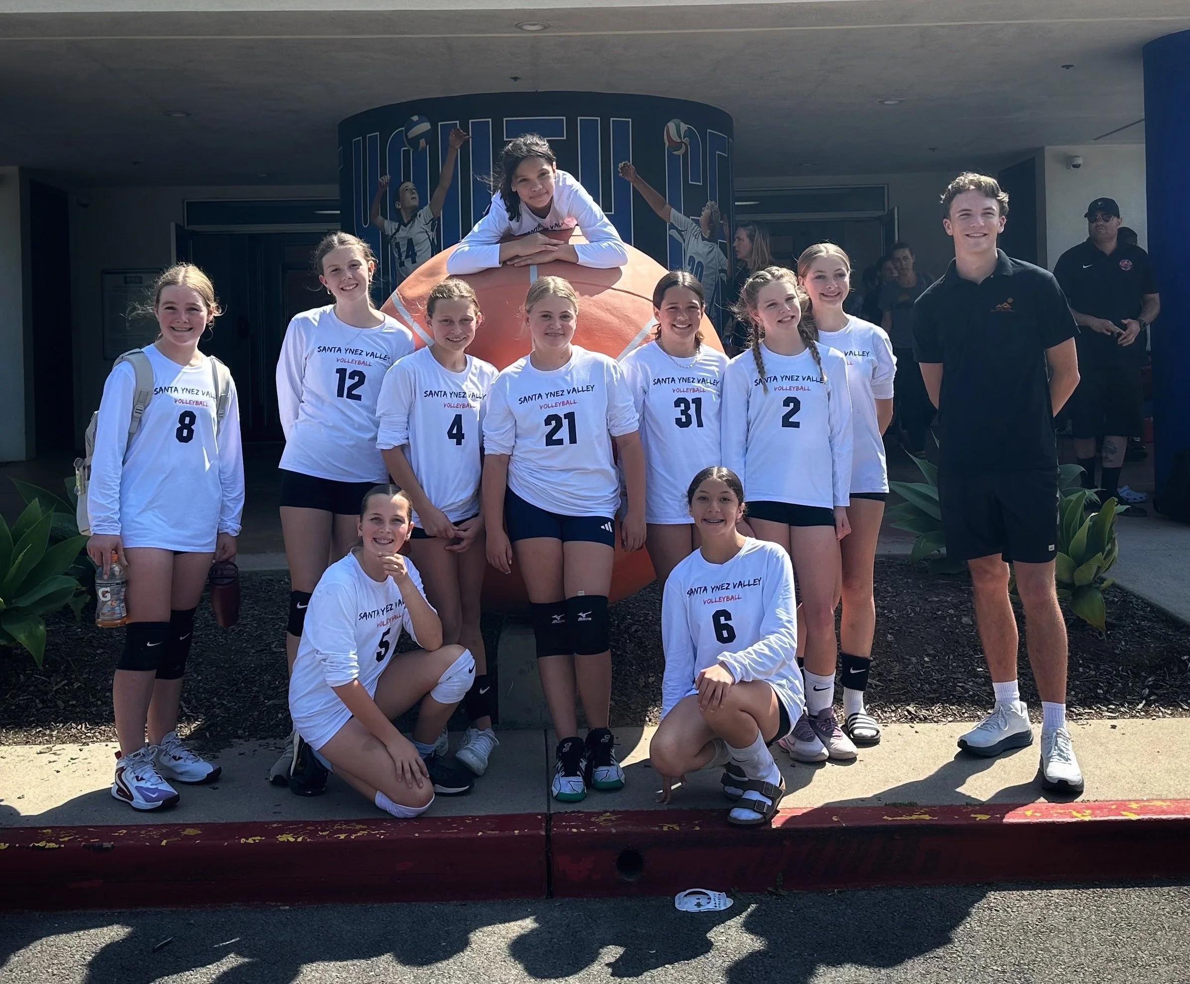 A girls' volleyball team from Santa Ynez Valley, posing in front of a large volleyball sculpture with their coach indoors at a school or sports facility.