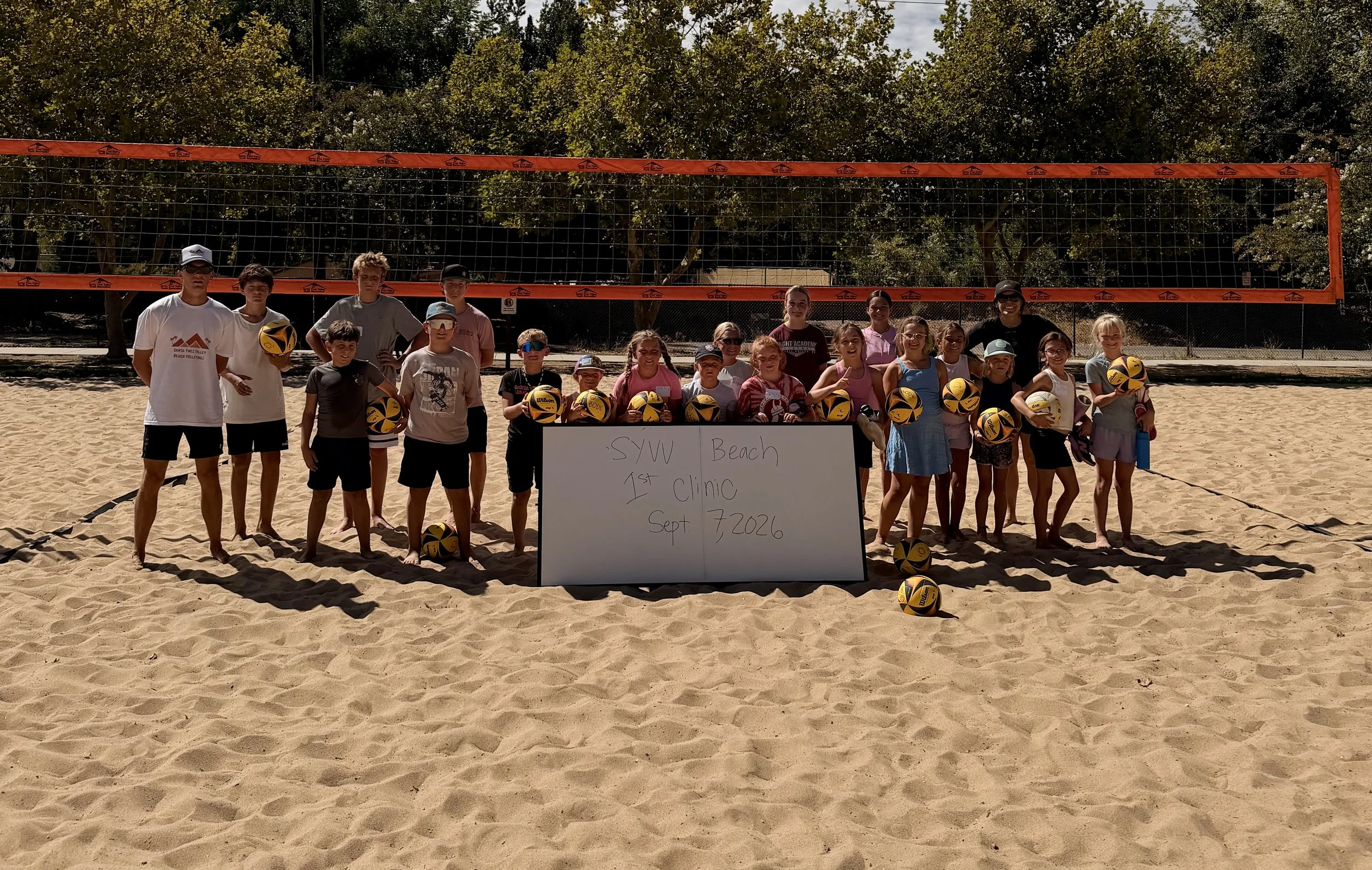 A group of children and two adults on a sandy beach beside a volleyball net. The group is holding volleyballs and standing behind a whiteboard with the text 'SWU Beach 1st Clinic Sept 7, 2026'. Trees are visible in the background.