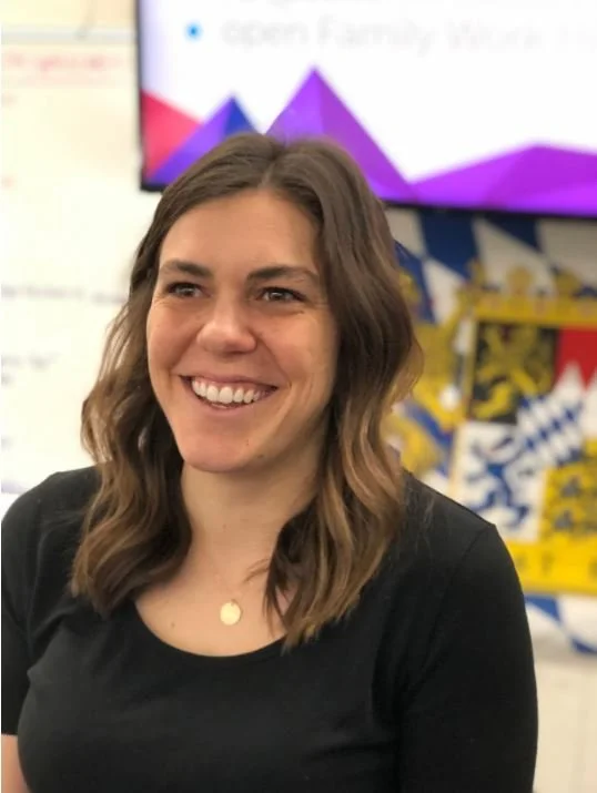 A smiling woman with brown, wavy hair wearing a black top, standing indoors with a colorful, geometric background and a screen in the background.