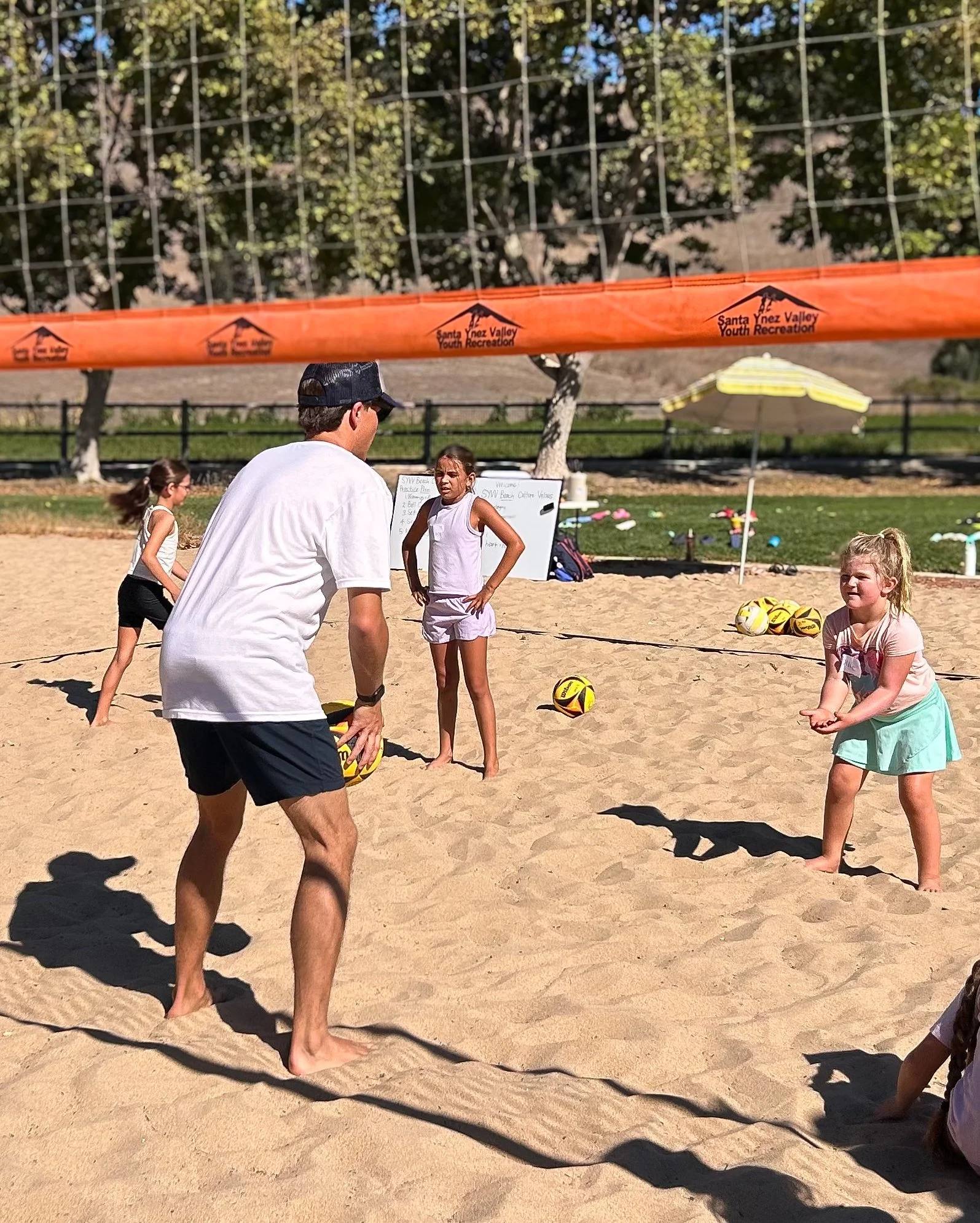 Children practicing volleyball on a sandy beach court, with a coach or instructor and a volleyball net, surrounded by trees and sunshine.