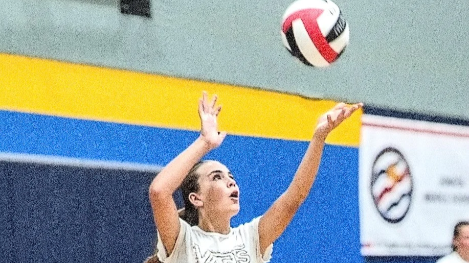 A young woman playing volleyball indoors, preparing to serve or pass the ball, with a colorful wall in the background.