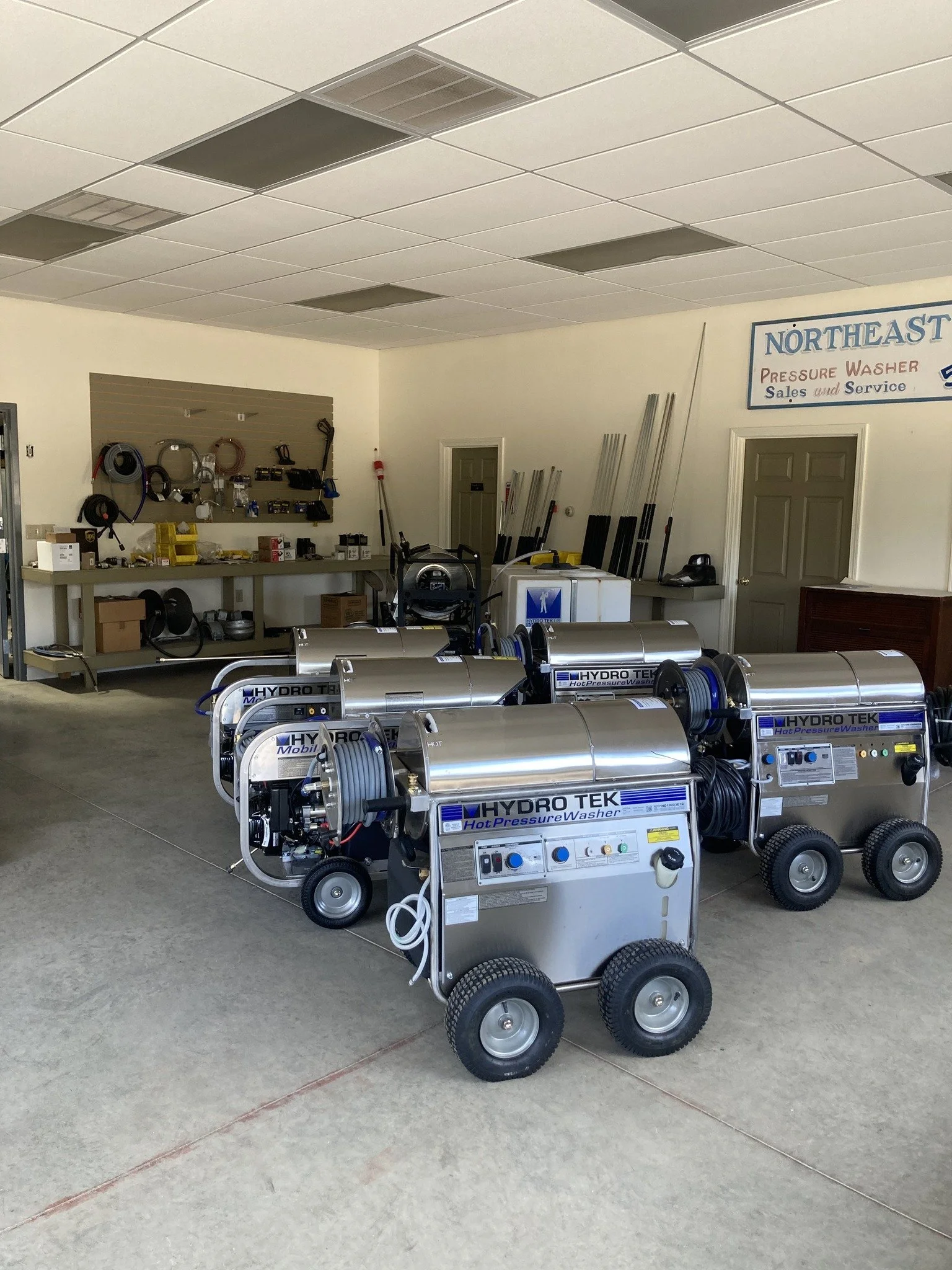 Three industrial pressure washers labeled 'HYDRO TEK Hot Pressure Washer' in front of a workspace with tools and equipment, inside a workshop or supply store.