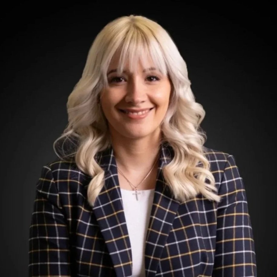 A young woman with long blonde hair and a cross necklace, smiling indoors with a large tree trunk in the background.