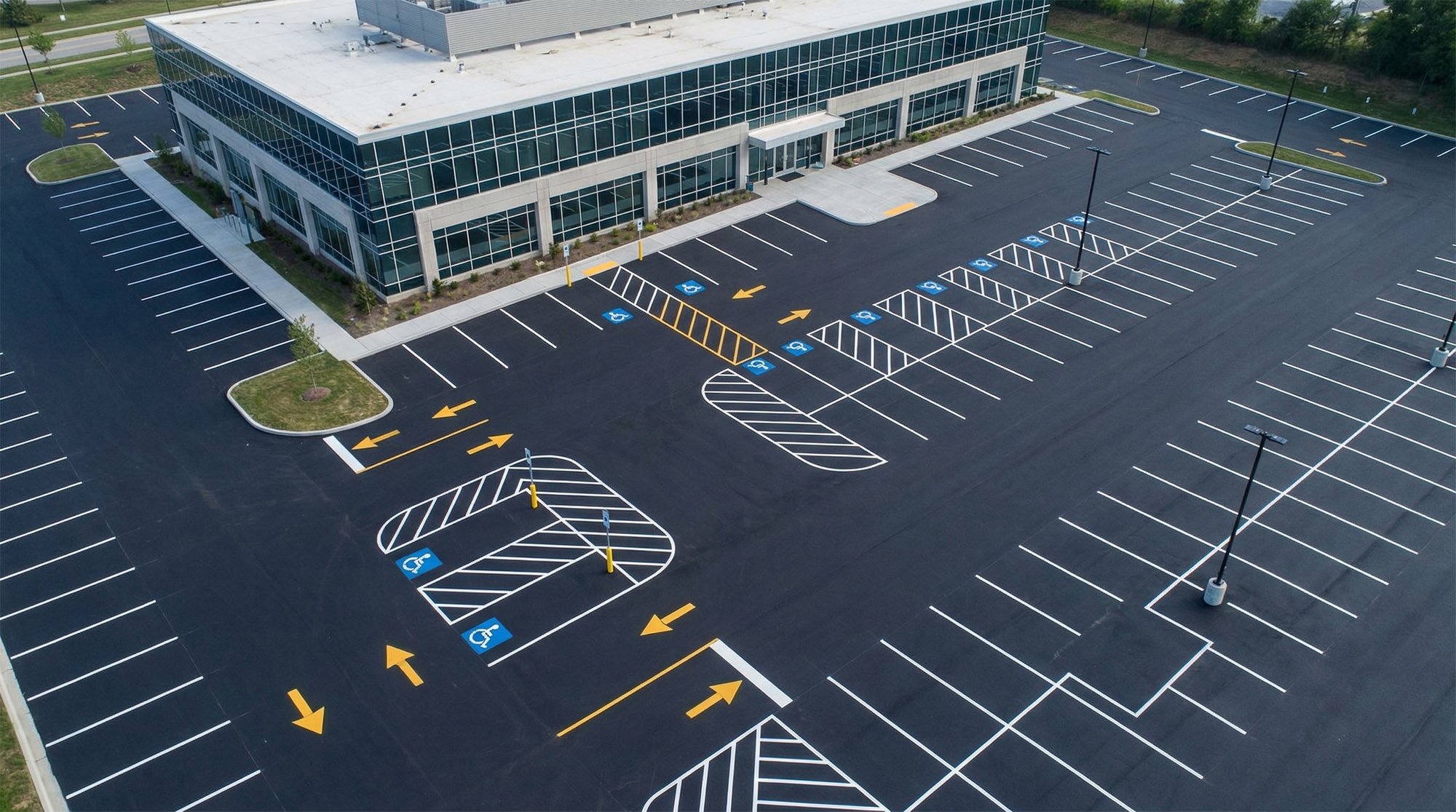 Empty parking lot with accessible parking spaces, yellow directional arrows, and a modern glass building in the background.