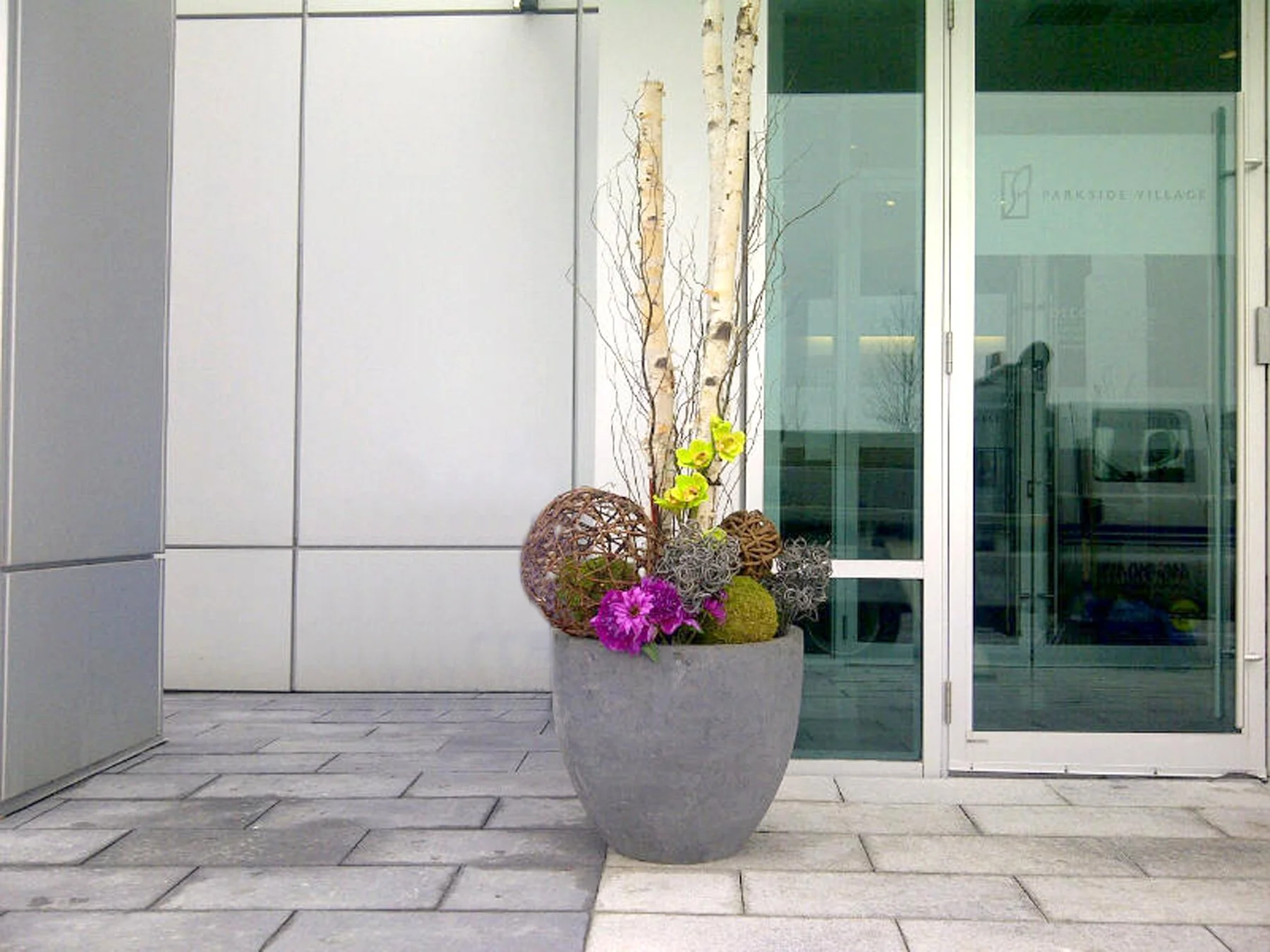 A large flower pot with a decorative arrangement of colorful flowers and ornamental balls placed outside beside a modern building's glass entrance door.