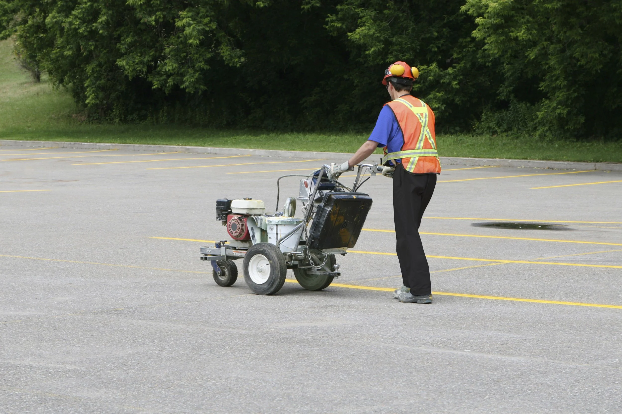 A worker standing in an empty parking lot, wearing a safety vest, helmet, and gloves, operating a line striping machine.