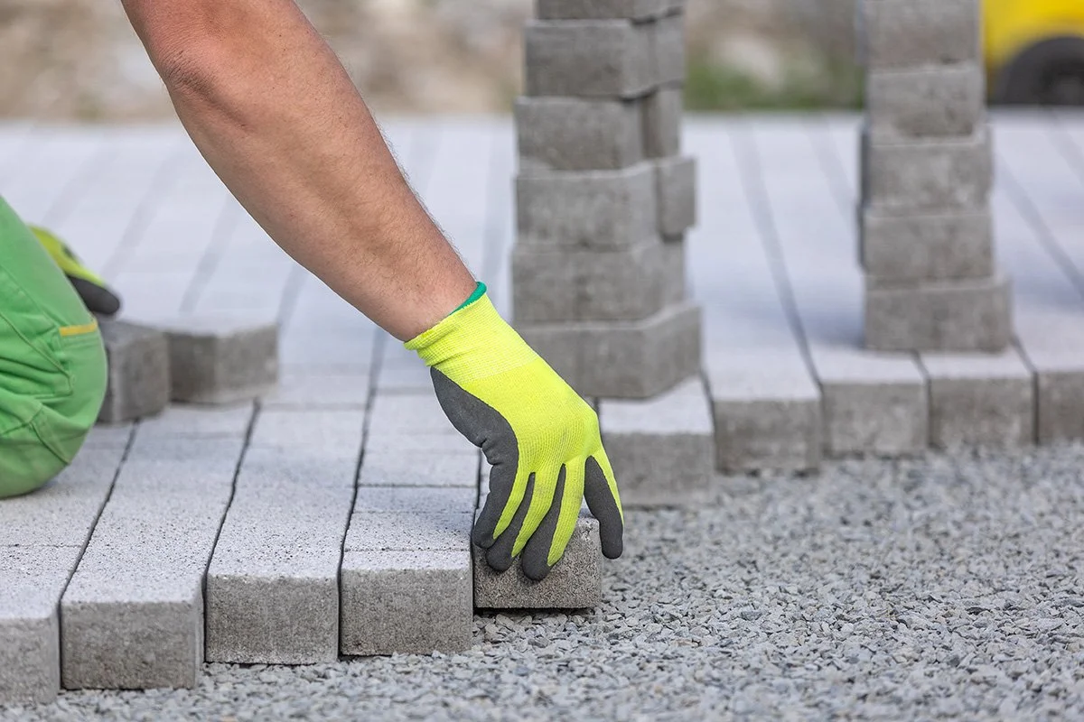 A person laying gray pavers in a pattern on a gravel surface, wearing yellow gloves.