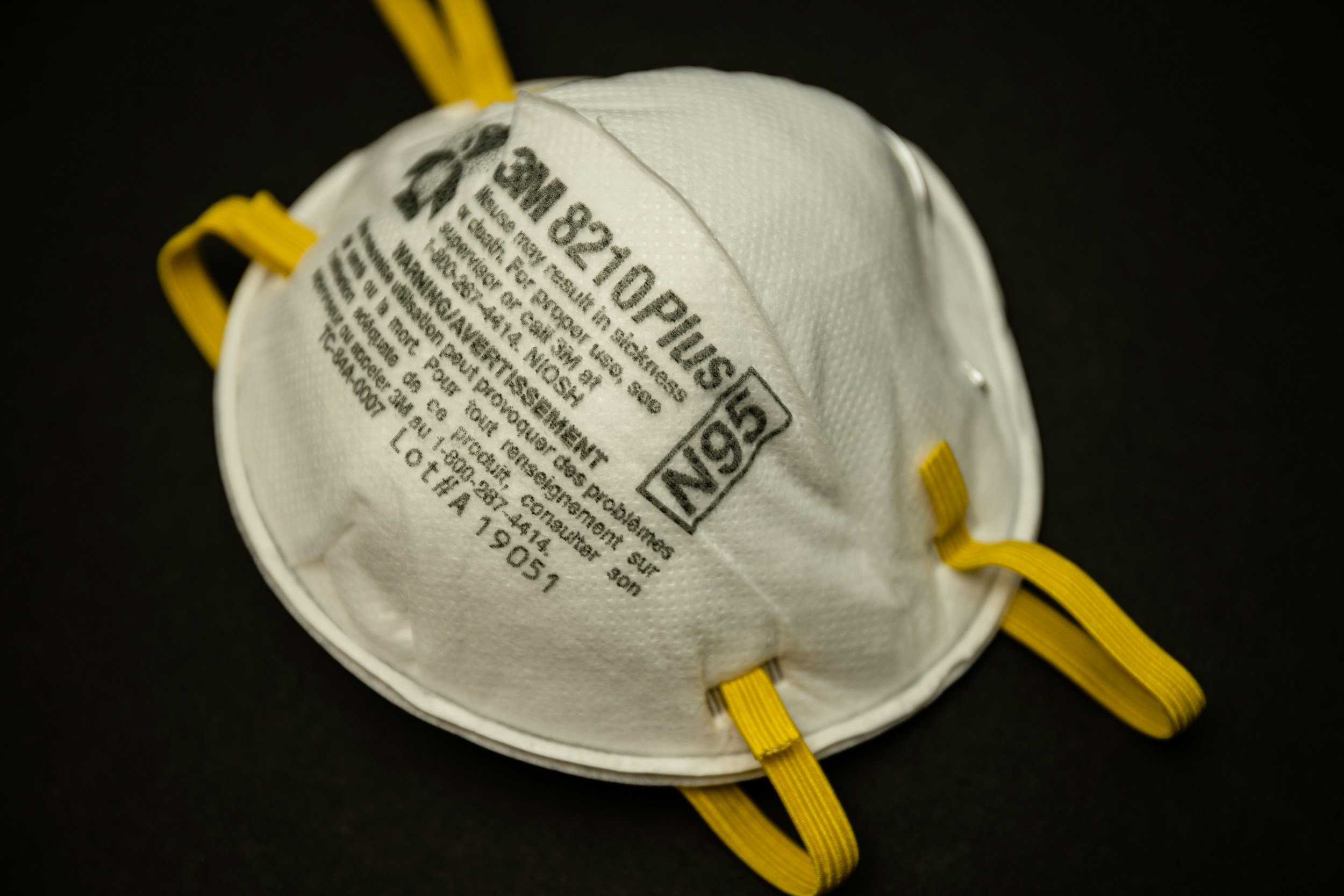 Close-up of a white N95 particulate respirator mask with yellow straps, against a black background.