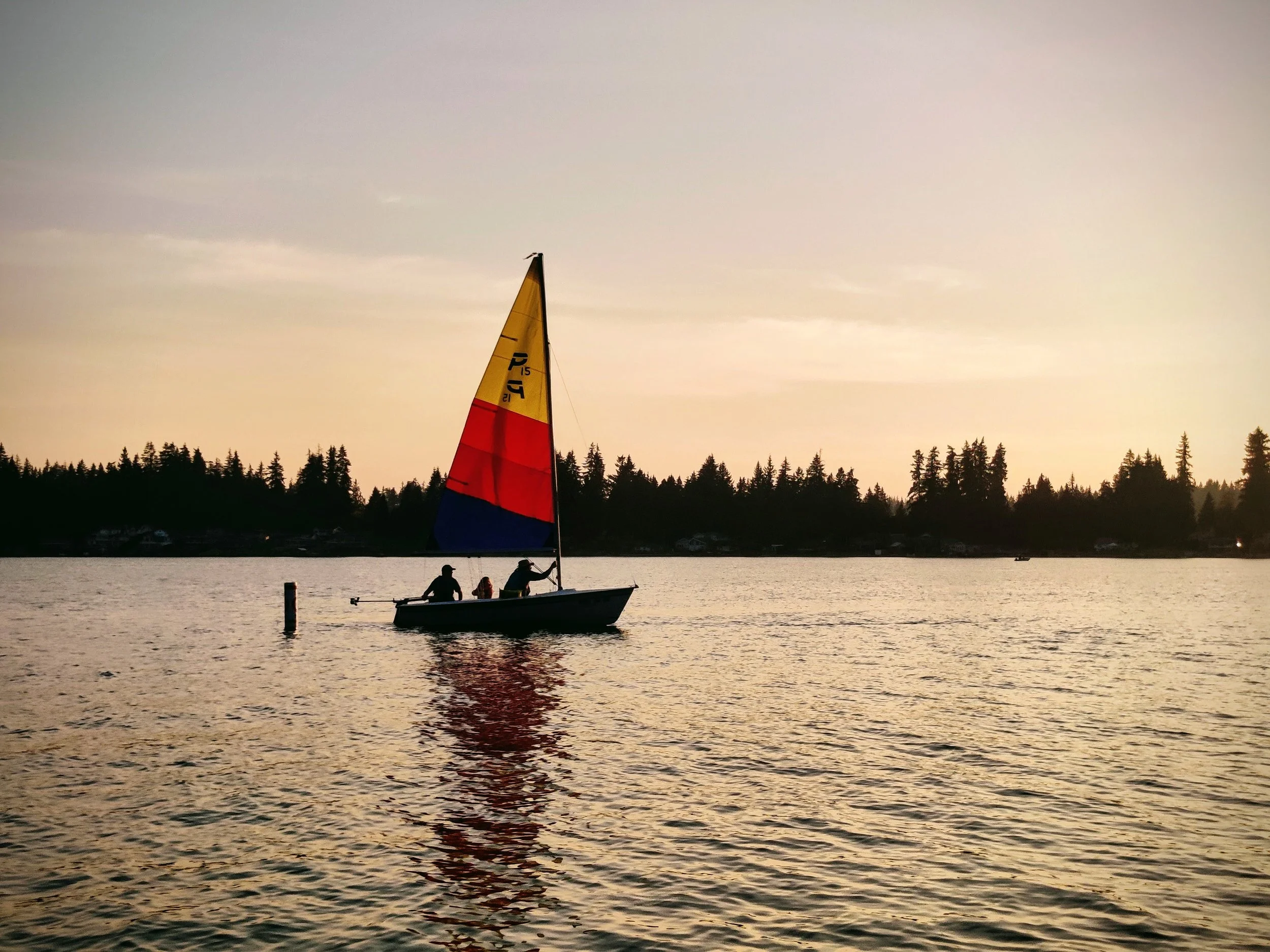 A sailboat with colorful sails on a calm body of water during sunset or sunrise, with a tree-lined shoreline in the background.