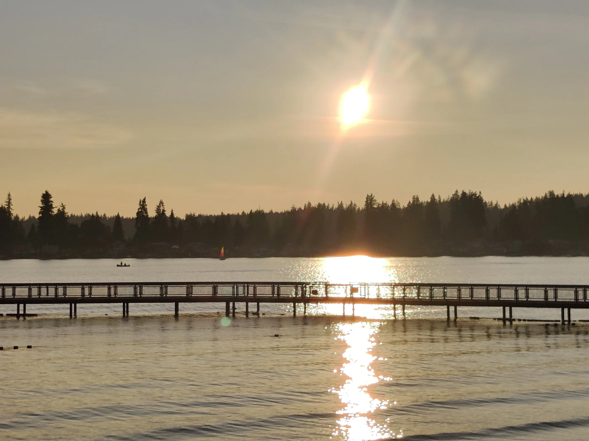 A sunset over a body of water with a wooden pier in the foreground, trees lining the horizon, and a small boat with two people and a sailboat visible in the distance.