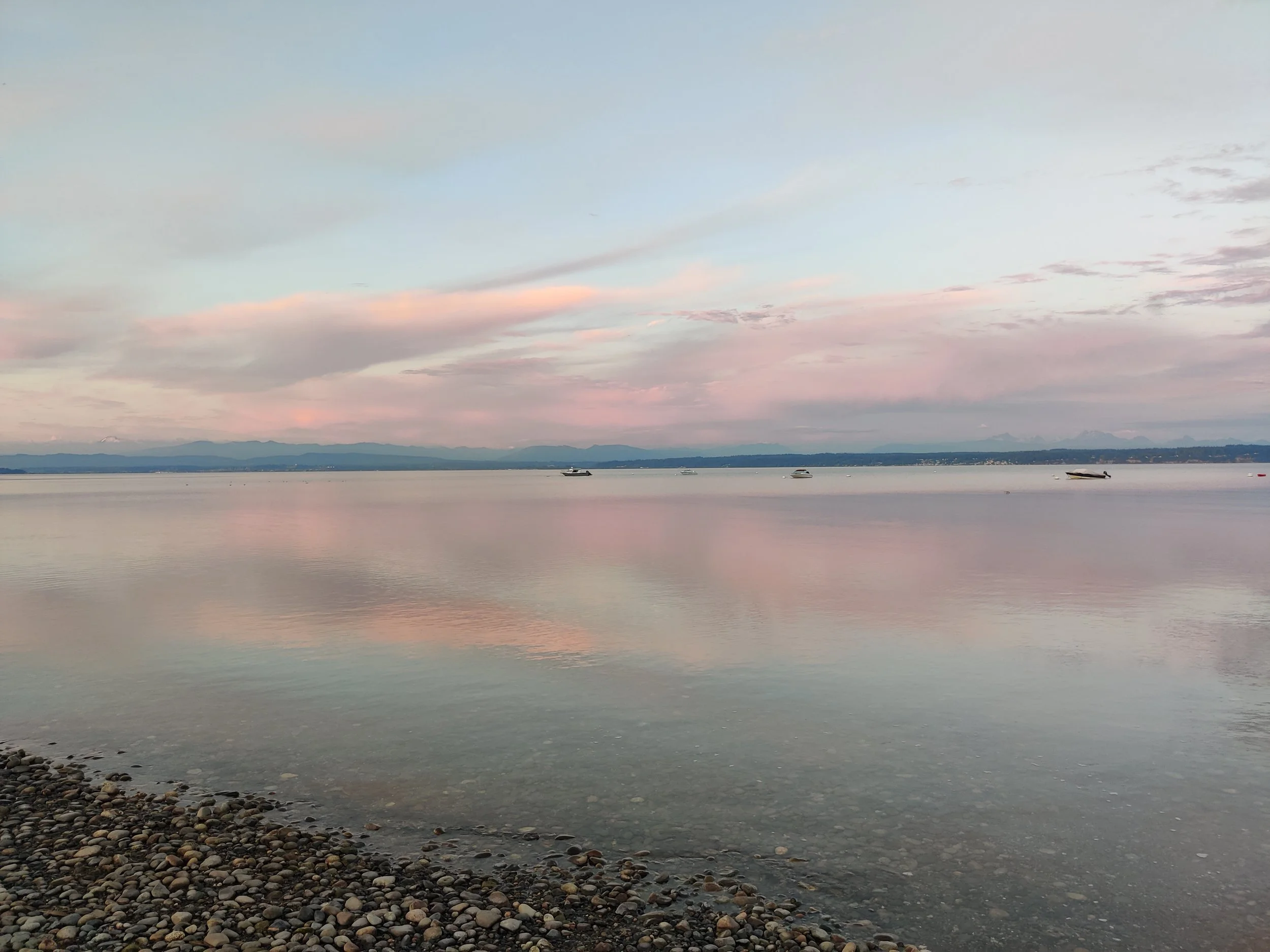 A calm body of water at sunset with a light pink and blue sky, distant mountains on the horizon, and several boats floating in the water near the shoreline of pebbles.