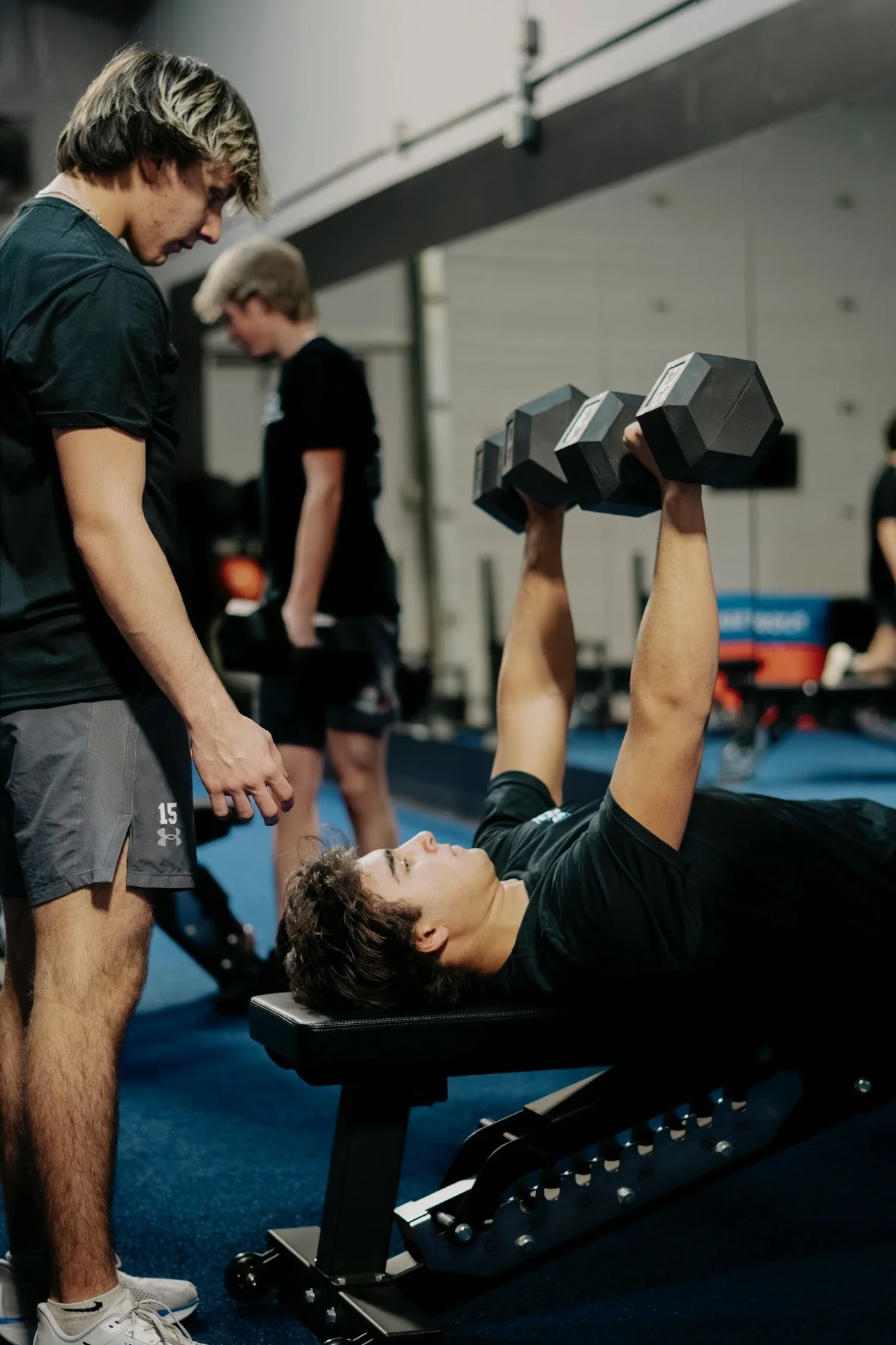 A young man lying on a workout bench, lifting dumbbells, while a trainer watches him in a gym.