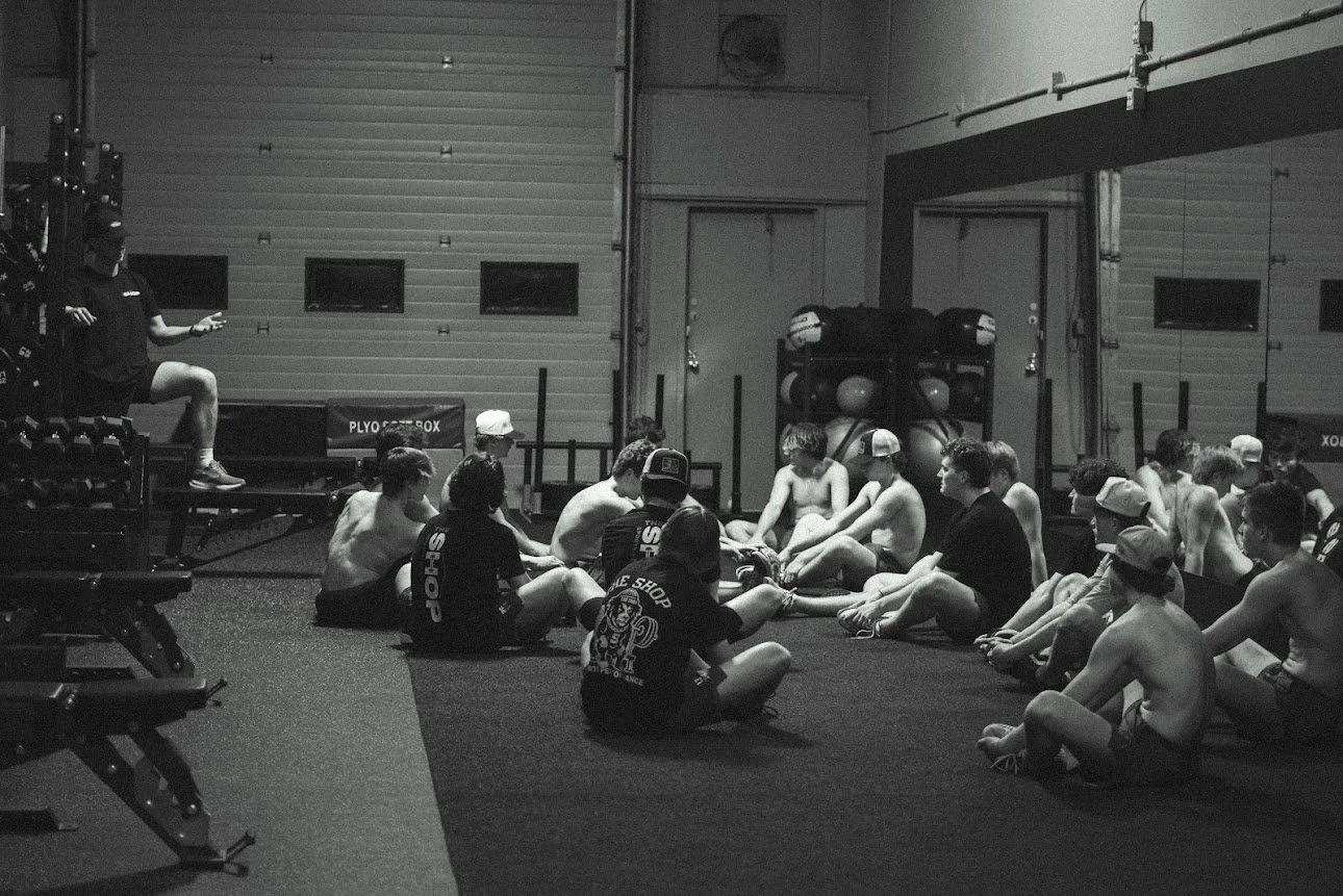 Group of young boys sitting on the gym floor, some shirtless, in a circle in what appears to be a fitness or martial arts gym. An instructor or coach stands on the left, demonstrating or explaining something to the group. Sporting equipment and gym mats are visible in the background.