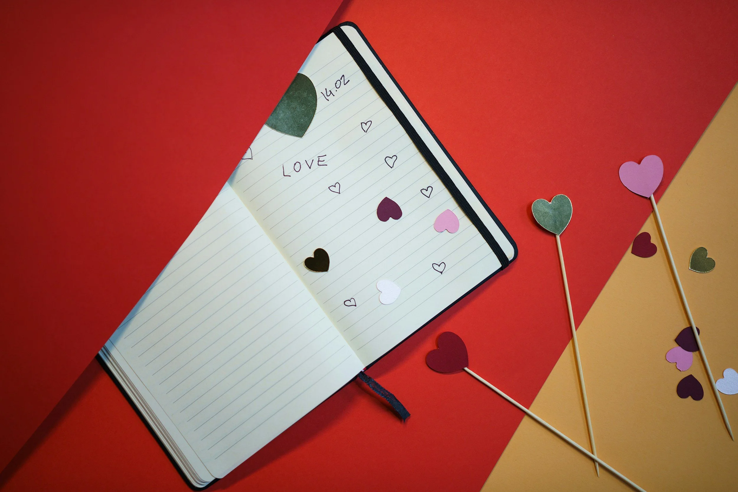 Open notebook with the word LOVE and handwritten hearts on the pages, set against multicolored red, yellow, and orange backgrounds, with paper heart confetti, some on toothpicks.