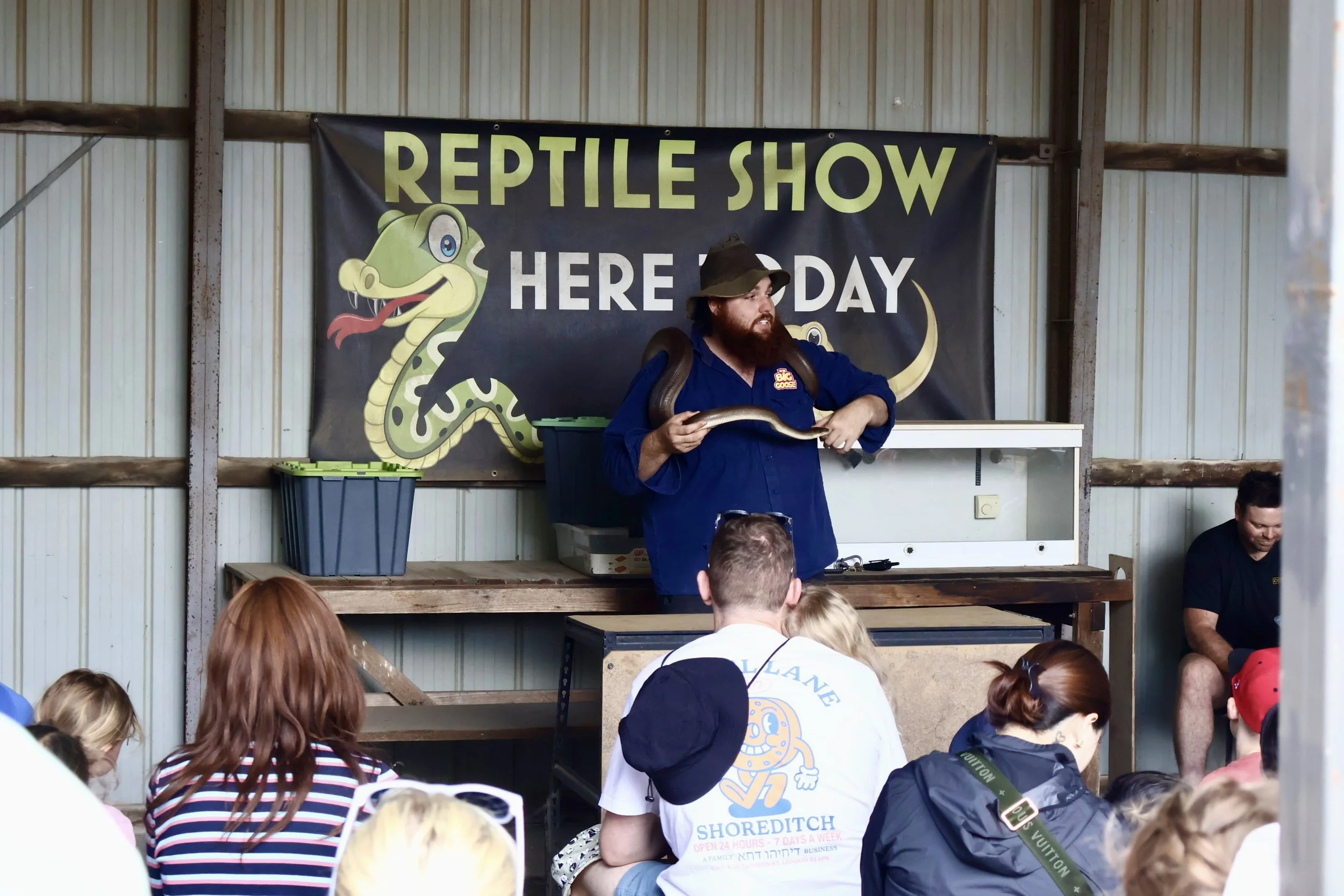 A reptile show event with a man holding a large snake in front of a banner that reads 'Reptile Show Here Today,' with a cartoon snake illustration, and an audience watching.