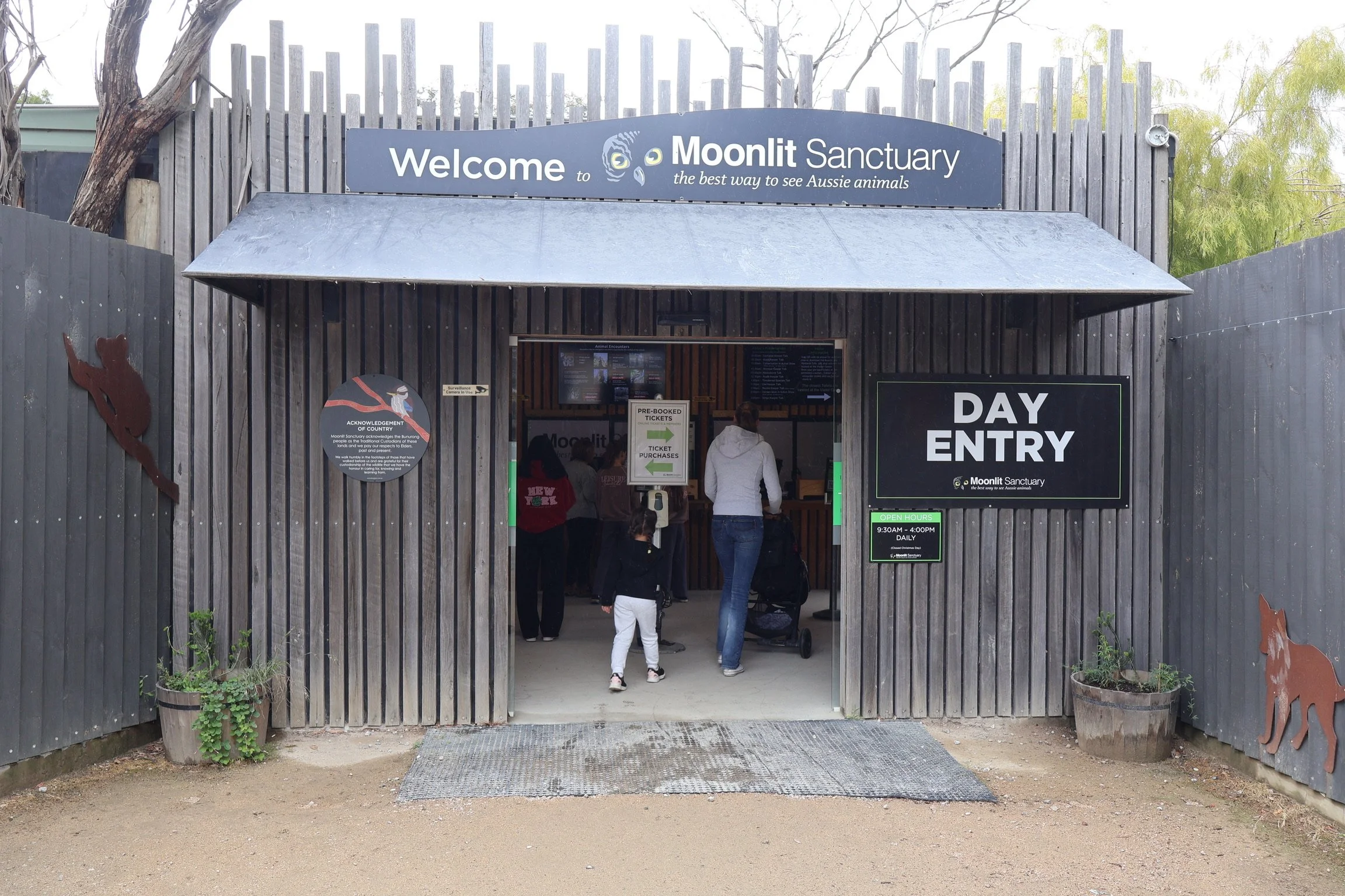 Entrance to Moonlit Sanctuary, an Australian animal sanctuary, with visitors entering and signs indicating day entry hours and ticket information.
