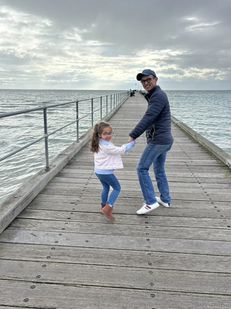 A man and young girl holding hands and smiling on a wooden pier extending over the water, with cloudy sky above.