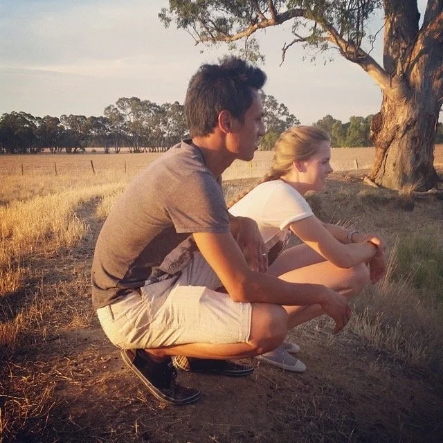 A young man and woman crouch on the grass in a rural setting, observing nature during sunset, with trees and open fields around them.